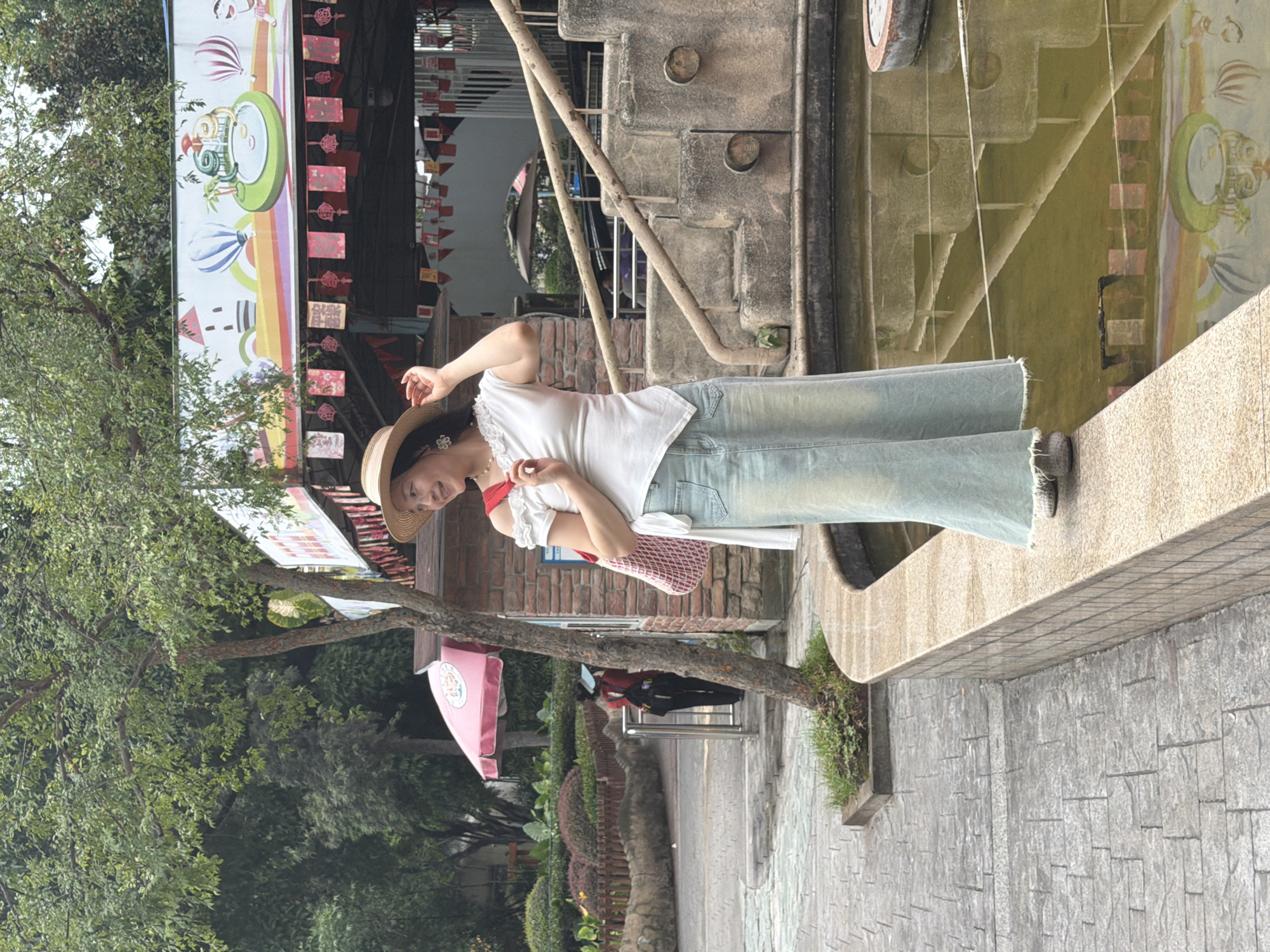 A woman wearing a white off-the-shoulder top, a light blue denim skirt, and a straw hat stands on the edge of a decorative fountain in what appears to be a park or outdoor recreational area. She is holding a red and white patterned bag and has her hand on her hat. The background shows a brick building, stairs with railings, and trees. There is a colorful mural visible in the reflection of the fountain water. In the distance, another person wearing a red shirt and a blue hat is visible walking. The paving in front of the fountain is made of stone tiles. This scene is located in Jiulongpo District, China.