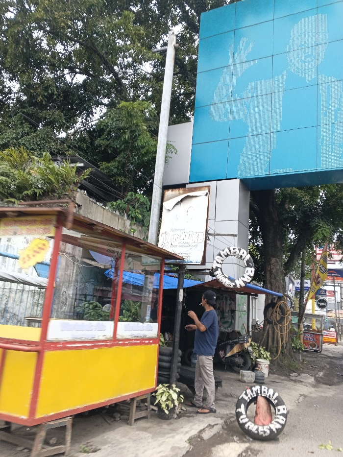 A street scene shows a yellow and red food cart next to a tire repair shop. A man wearing a blue shirt, tan pants, and a black cap stands by the tire repair shop with what appears to be a cigarette in his hand. Tires painted with "Tambal Ban" (tire repair) are used as signs. A large blue sign with a white image hangs above. Lush green trees are visible in the background.