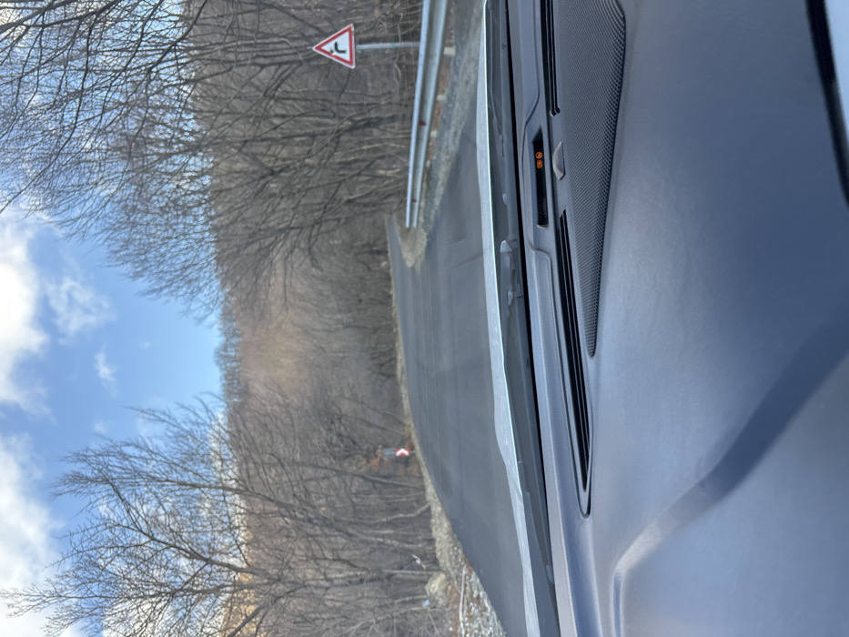 From inside a car, a winding road is seen through the passenger window. Leafless trees line the roadside under a blue sky with scattered clouds. A road sign indicating a bend is visible ahead. The car's interior, including the door panel, is partially in view.