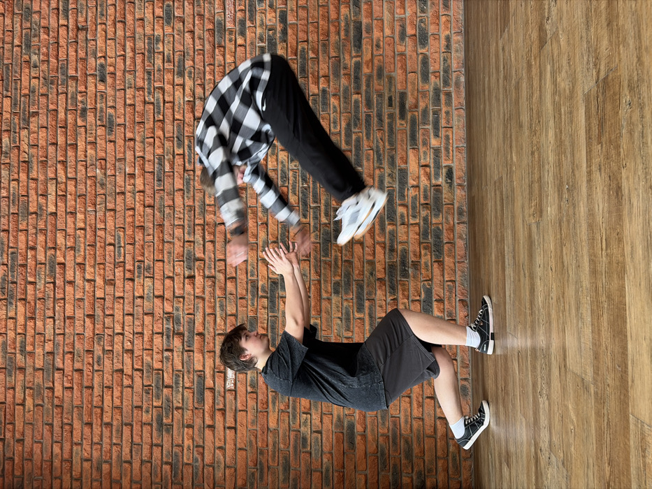 The image depicts an indoor scene in Chaikovsky, Russia, featuring two individuals engaged in a dynamic physical interaction against a backdrop of an orange-red brick wall and a light brown wooden floor.
On the left, a male individual with short dark hair, wearing a dark grey t-shirt, dark shorts, white socks, and black low-top sneakers, is positioned in a crouched stance, facing right. His knees are bent, and his arms are extended forward at chest height, palms open, appearing to brace or push. His gaze is directed towards the second individual.
On the right, a second male individual, largely obscured by motion blur, is airborne. He is wearing a black and white plaid long-sleeved shirt, dark pants, and white athletic shoes with distinct soles. His body is bent forward and slightly to the left, with both arms extended towards the hands of the first individual. His feet are off the ground, indicating a jump or propelled movement. The blurring suggests rapid motion.
The interaction shows the airborne individual moving towards or away from the stationary individual's extended hands, suggesting a coordinated physical activity, possibly a stunt, dance move, or active play. The environment is well-lit, providing clear visibility of the subjects and setting.