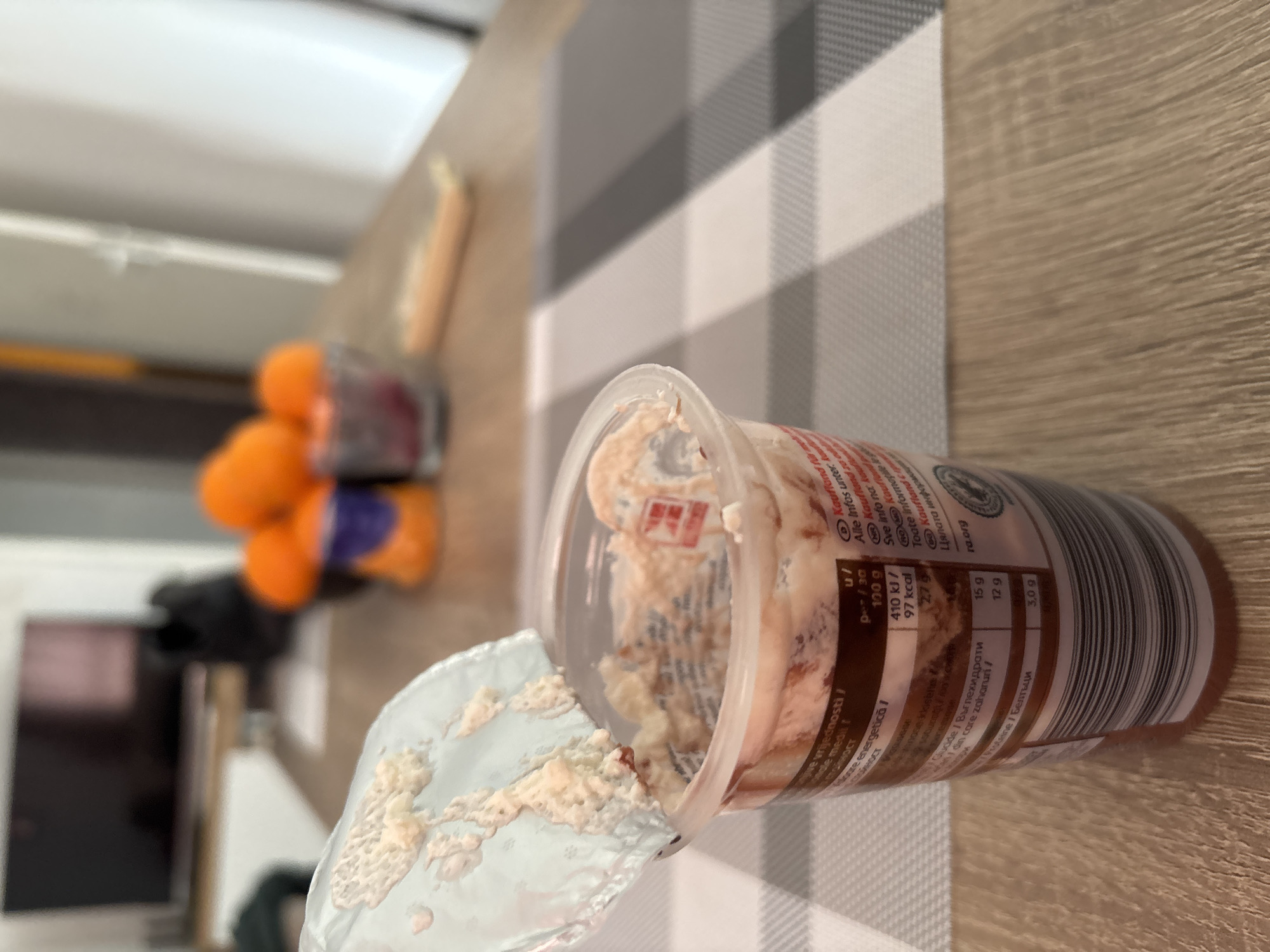 The image shows an opened container of what appears to be a spreadable cheese or dip, with the lid partially peeled back. The container sits on a table with a gray and white striped placemat. The nutrition information is visible on the container, with details in multiple languages including Bulgarian. In the background, there are blurred objects including what looks like oranges in a bowl and a dark object that could be a television or monitor. There is not enough information to determine if the scene is in Plovdiv, Bulgaria.