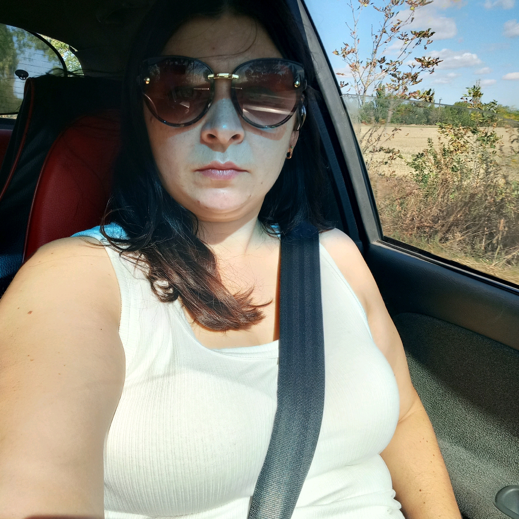 A medium shot captures a woman sitting in the passenger seat of a vehicle, looking directly at the camera. She is wearing a white tank top and large, dark sunglasses. A black seatbelt is fastened across her chest. Her dark hair falls around her shoulders. The interior of the car, including a red seat, is visible. Through the window, a sunlit rural landscape with trees and fields can be seen under a partly cloudy blue sky. The location is identified as Klyntsi, Ukraine.