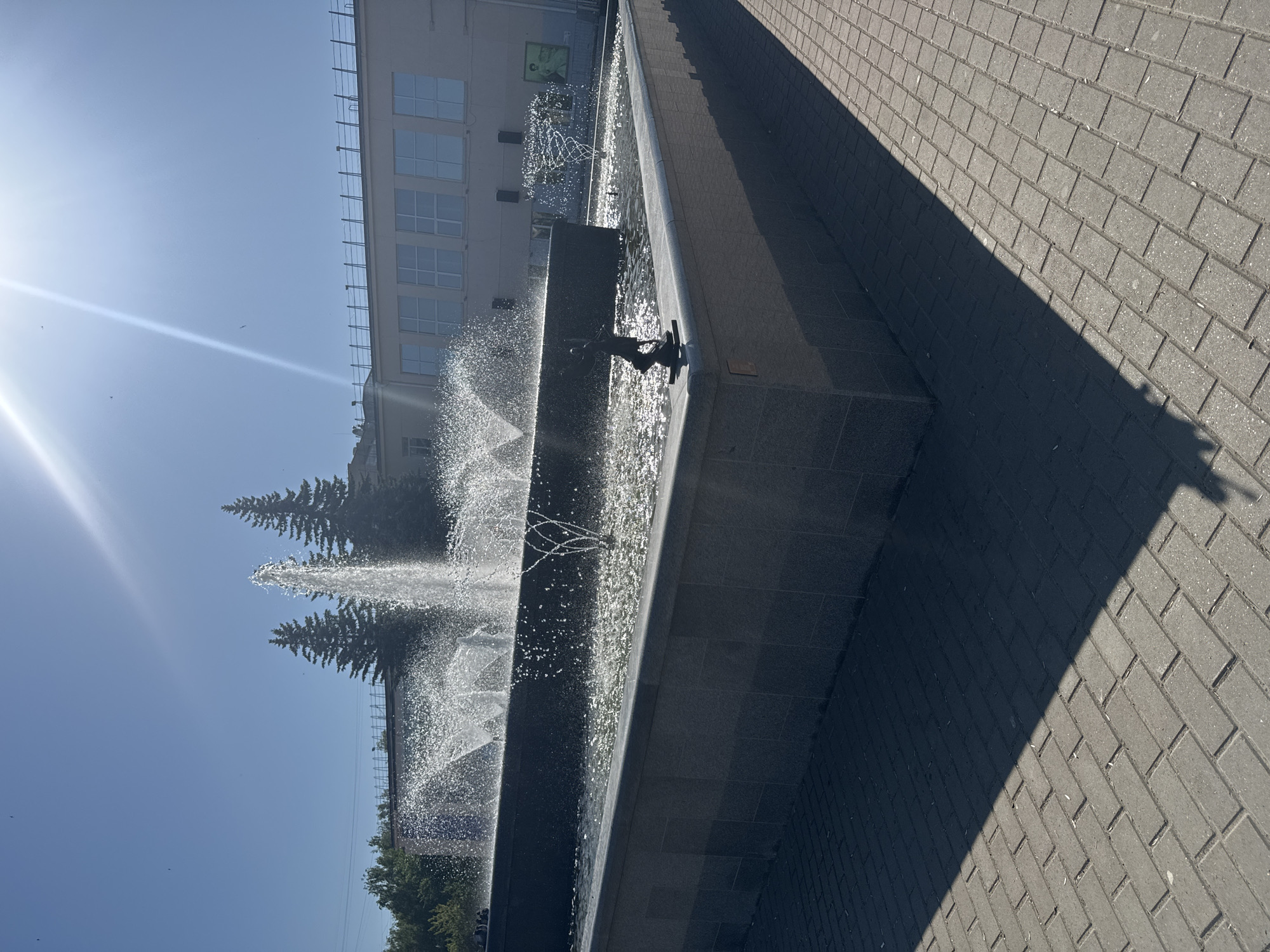 The image shows a fountain in the city of Chaikovsky, Russia. The fountain is a multi-tiered, rectangular structure made of dark gray stone. Water jets are visible, creating a dynamic display. Behind the fountain are evergreen trees and a multi-story building with several windows. To the right of the fountain is a brick-paved walkway, partially in shadow. The sky is bright blue, with the sun visible in the upper left corner. There are a few birds in the sky.