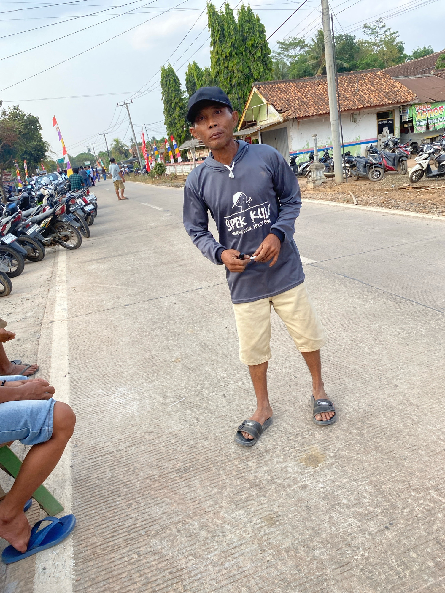 An Indonesian man wearing a dark gray hoodie, khaki shorts, a black baseball cap, and dark gray slides stands on a concrete road. He is holding a cigarette in his right hand. The road is lined with parked motorcycles and various buildings. In the background, flags are visible, suggesting a possible event or celebration. The sky is overcast, and the overall lighting suggests it is late afternoon or early evening. The location is identified as Sri Sawahan, Indonesia.