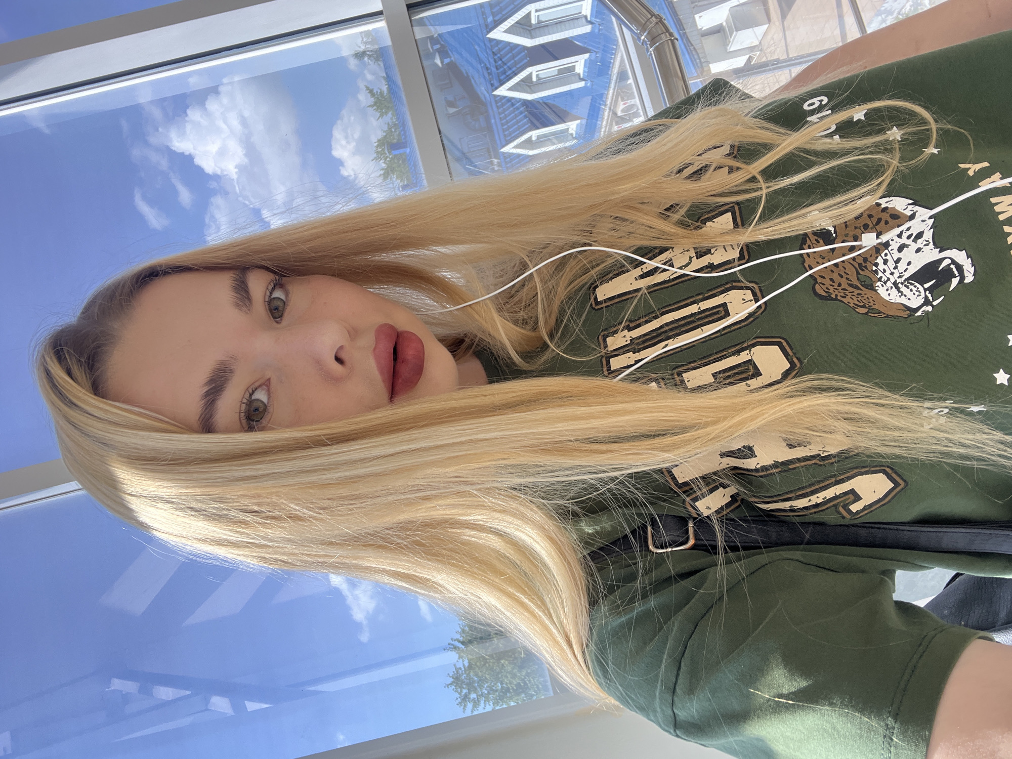 This is a selfie of a young woman with long blonde hair, wearing a green t-shirt with a graphic of a leopard and text. She is also wearing white earbuds. The background appears to be an indoor space with large windows, offering a view of a blue sky with clouds and some buildings. It is daytime, and the lighting suggests the sun is shining. The reflection on the windows indicates an urban environment. The woman is looking directly at the camera.