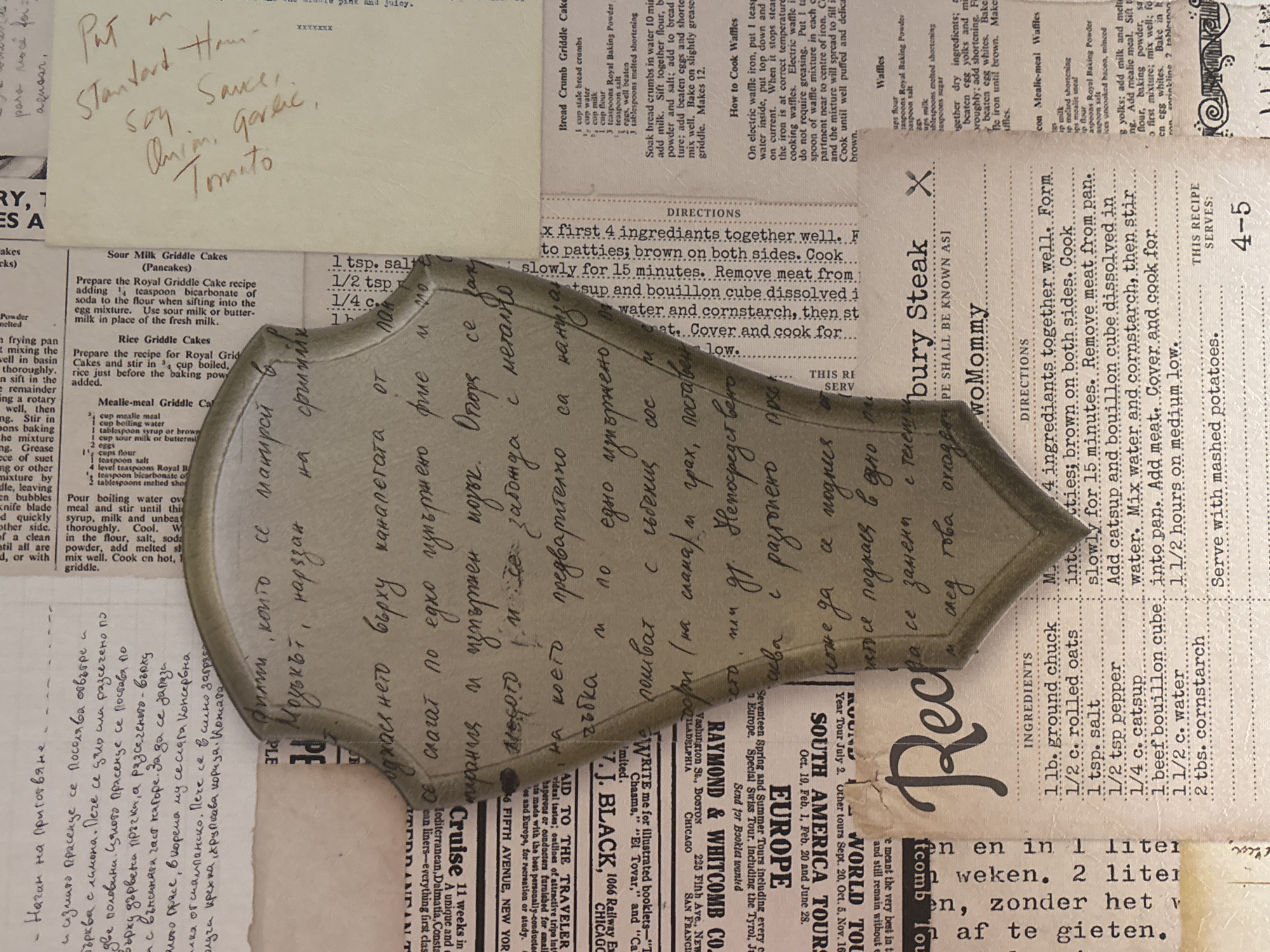 This image shows a collection of documents and objects. There are several pieces of paper covered with text, including recipe cards and advertisements, in both English and another language (possibly Bulgarian). There is a handwritten note at the top left reading "Put on Standard Ham-sauce: onion, garlic, tomato." A metal shield shape is placed in the center, covering part of the other texts. The overall composition suggests a collage or a collection of miscellaneous papers arranged on a surface.