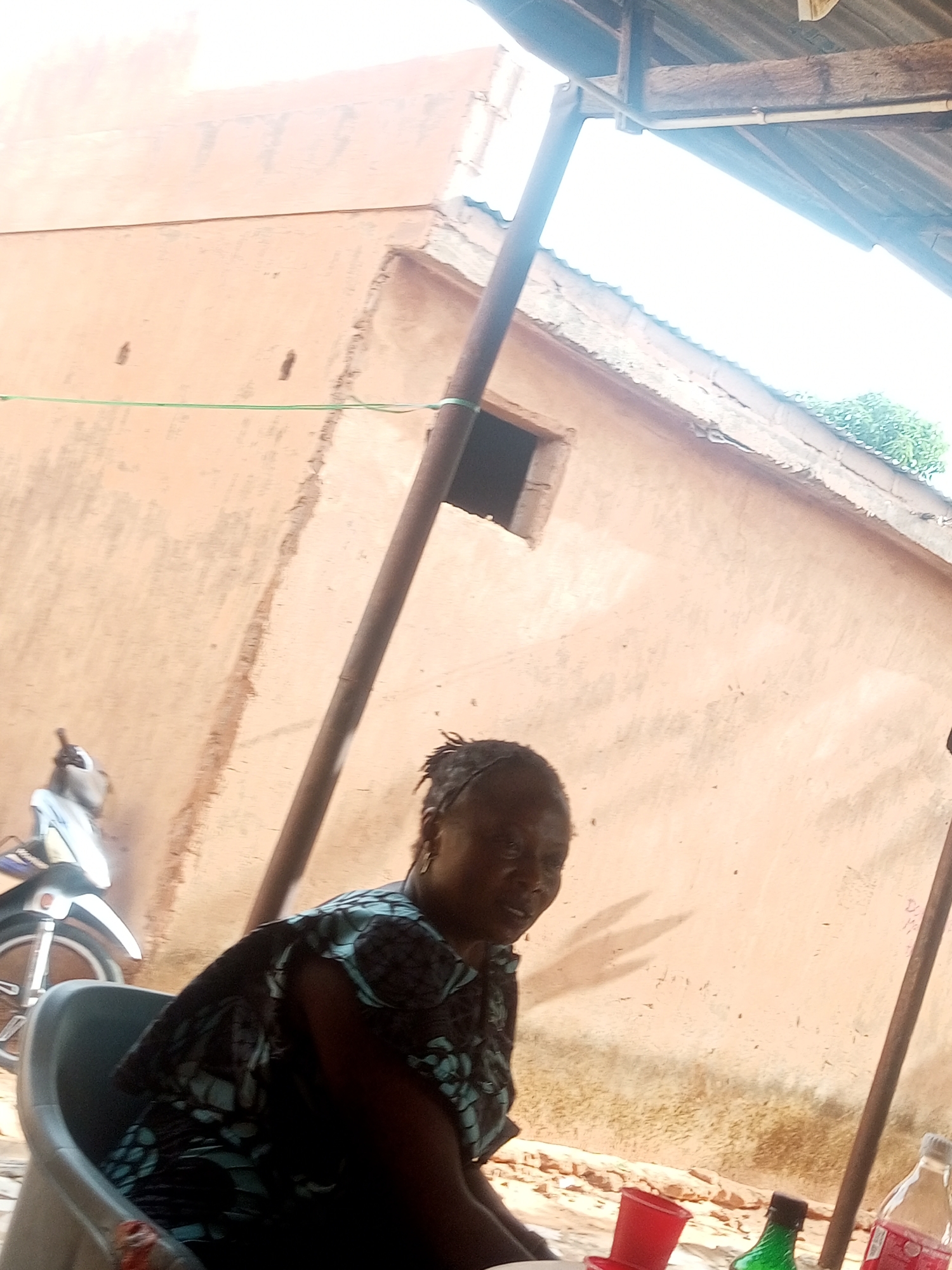 The image shows an unidentified person sitting in a gray plastic chair, possibly outdoors, in the city of Unknown, Unknown. In the background is a peach-colored building with a small window, and a partial view of a motorcycle on the left. A green string is stretched horizontally across the frame. A rusty metal post is positioned on the right side of the frame, and several bottles and red plastic cups sit on a surface next to the individual. The area seems to be covered by a roof made of corrugated metal.