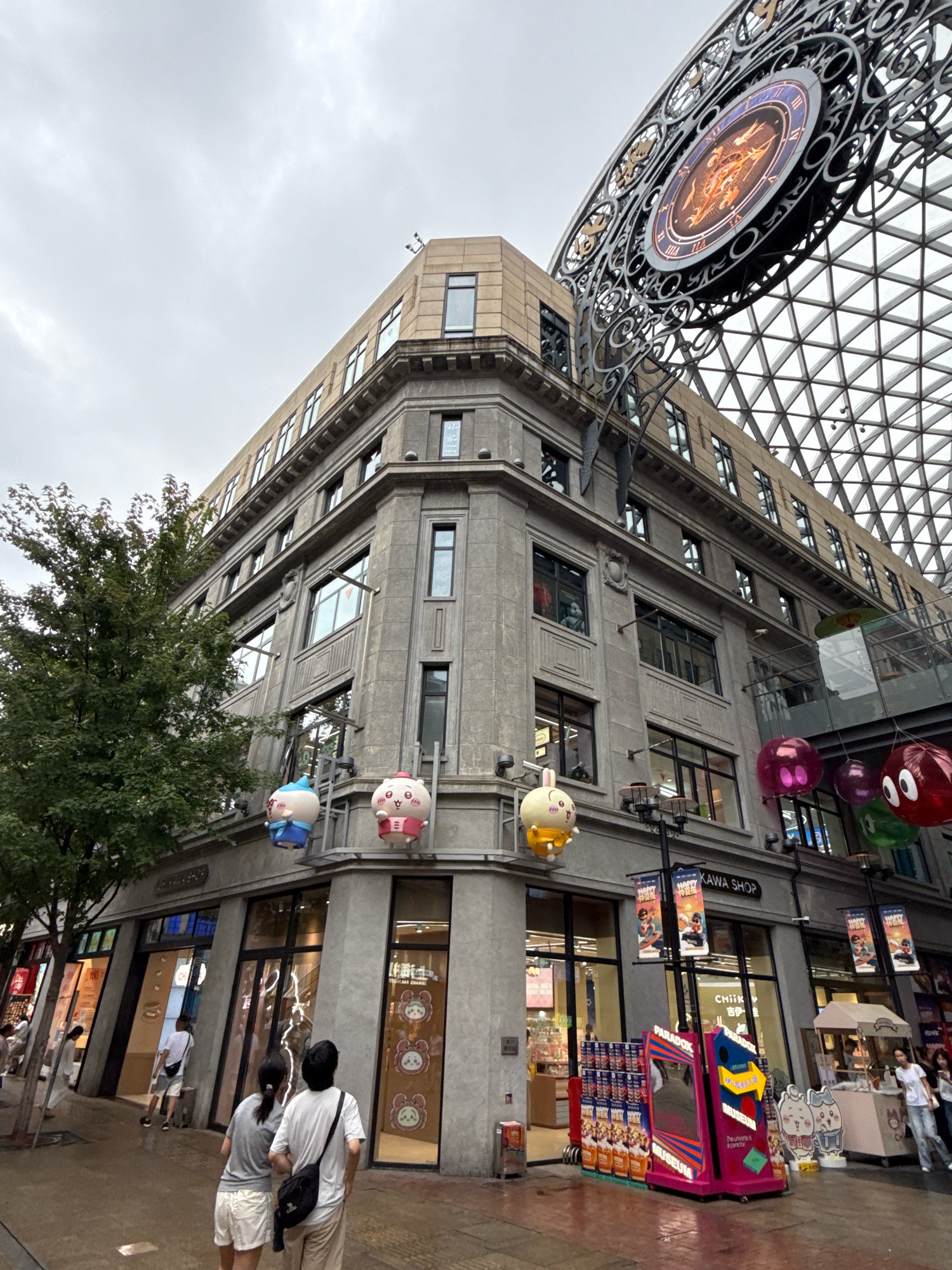 The image captures a street view in Shanghai, China, likely within a shopping district, under an overcast sky. A multi-story building with a stone facade occupies the center and right side of the frame. The building features numerous windows and decorative architectural elements. Hanging from the upper floors are large, inflatable cartoon character balloons in various colors, including pink, yellow, and blue. A prominent circular clock with ornate metalwork is visible in the upper right corner, part of a larger structure that appears to be a geodesic dome or similar roof.
At street level, storefronts with large glass windows line the building. One sign reads "KAWA SHOP". In front of one of the shops, a display includes colorful boxes and a pink, multifaceted structure with the word "PARADOX MUSEUM" written on it. Several people are present. Two individuals, a woman and a man, are walking away from the camera, with the woman wearing a grey t-shirt and shorts and the man wearing a white t-shirt and light-colored pants. They both have bags slung over their shoulders. Other individuals are seen walking in the background, and a food cart is present near the right edge of the frame. A tree with green foliage stands to the left of the building. The ground appears to be wet, suggesting recent rain.