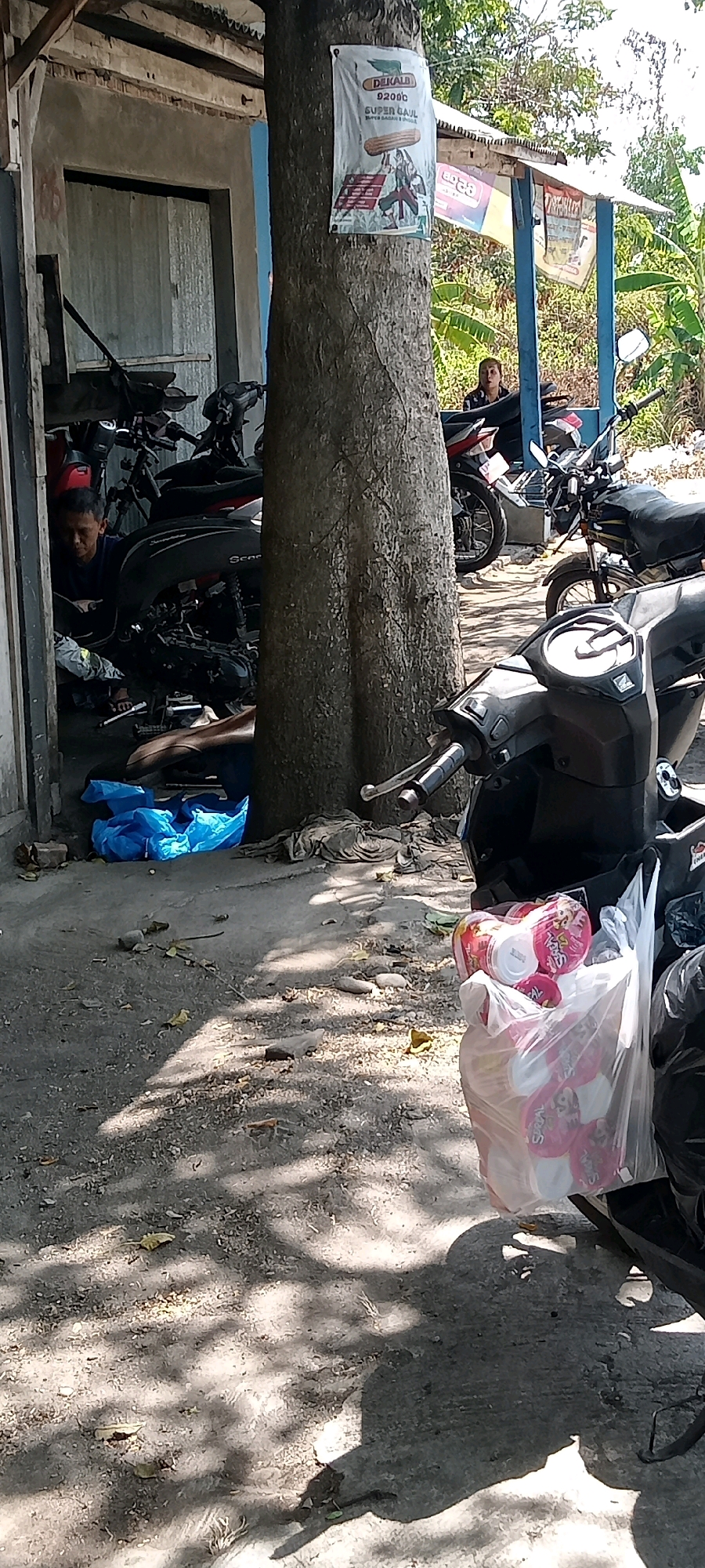 The image captures a street scene in Kemusu, Indonesia. In the foreground, a black motorcycle is parked, with a clear plastic bag filled with items, possibly snacks or toiletries, hanging from its handlebars. The bag is transparent and the contents are visible. Behind the motorcycle, a large tree trunk stands prominently. To the left, a small workshop-like structure is visible, with a man working on a motorcycle inside. Several other motorcycles are parked in and around the workshop. Further in the background, another motorcycle is parked under a blue awning, and a person is seated on it. The environment appears to be outdoors, with a dirt and concrete ground, littered with some leaves and debris. Sunlight casts shadows on the ground, indicating it is daytime. A poster is affixed to the tree trunk, featuring text and images.