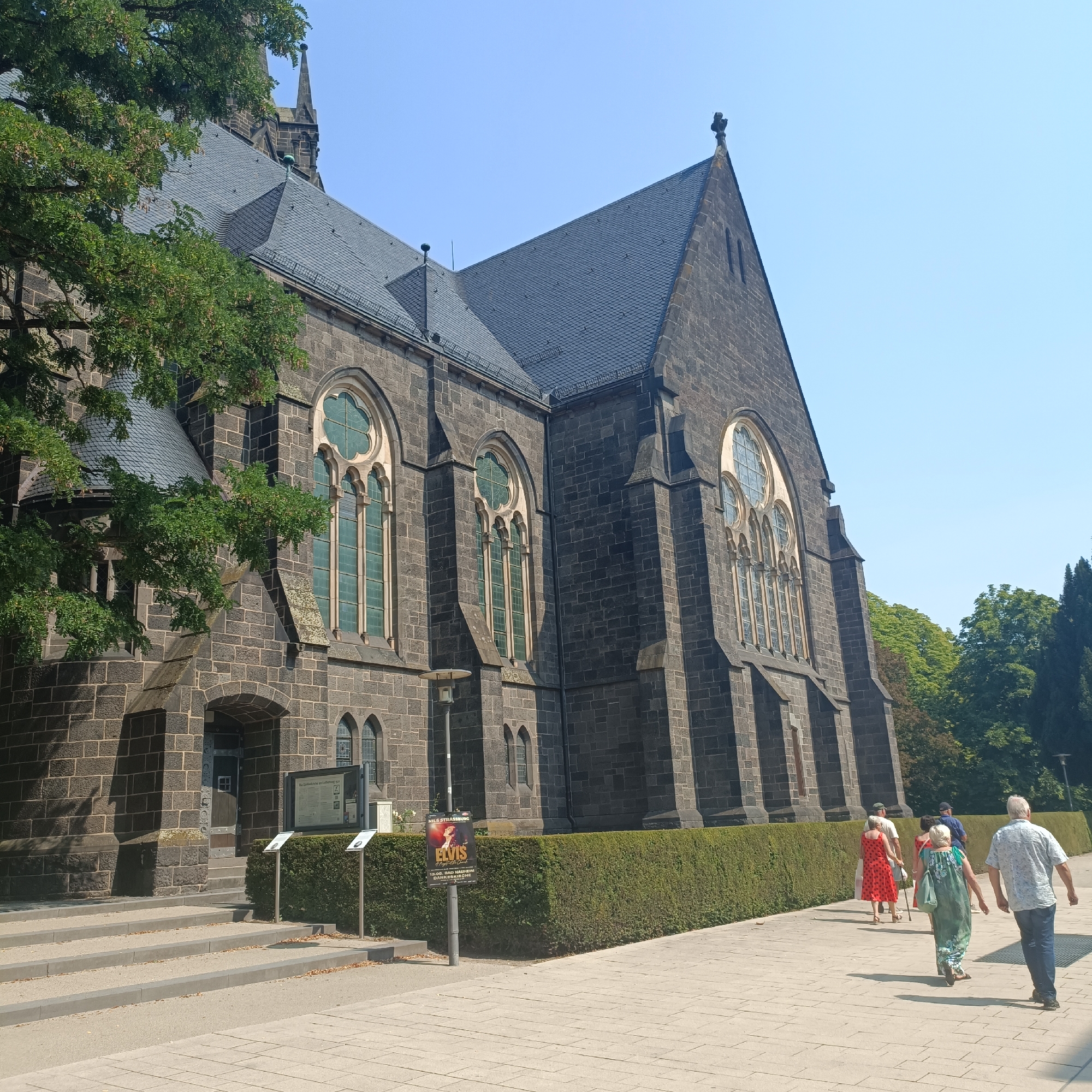 The image shows the exterior of the Dankeskirche (Church of Thanksgiving) in Bad Nauheim, Germany. The church is constructed of dark stone blocks and features a steeply pitched slate roof with a prominent cross at its apex. Multiple large, arched windows with intricate tracery and green-tinted glass are visible along the side of the building. A mature tree with green foliage partially obscures the left side of the church. In the foreground, a paved walkway extends alongside a manicured hedge. Several individuals are walking away from the viewer on the walkway. One woman in a red dress and a woman in a green patterned dress are particularly noticeable. Behind the hedge, a poster advertising an "Elvis" performance at the Dankeskirche is affixed to a pole. The sky above is clear and blue. The scene appears to be a sunny day, likely during the daytime.