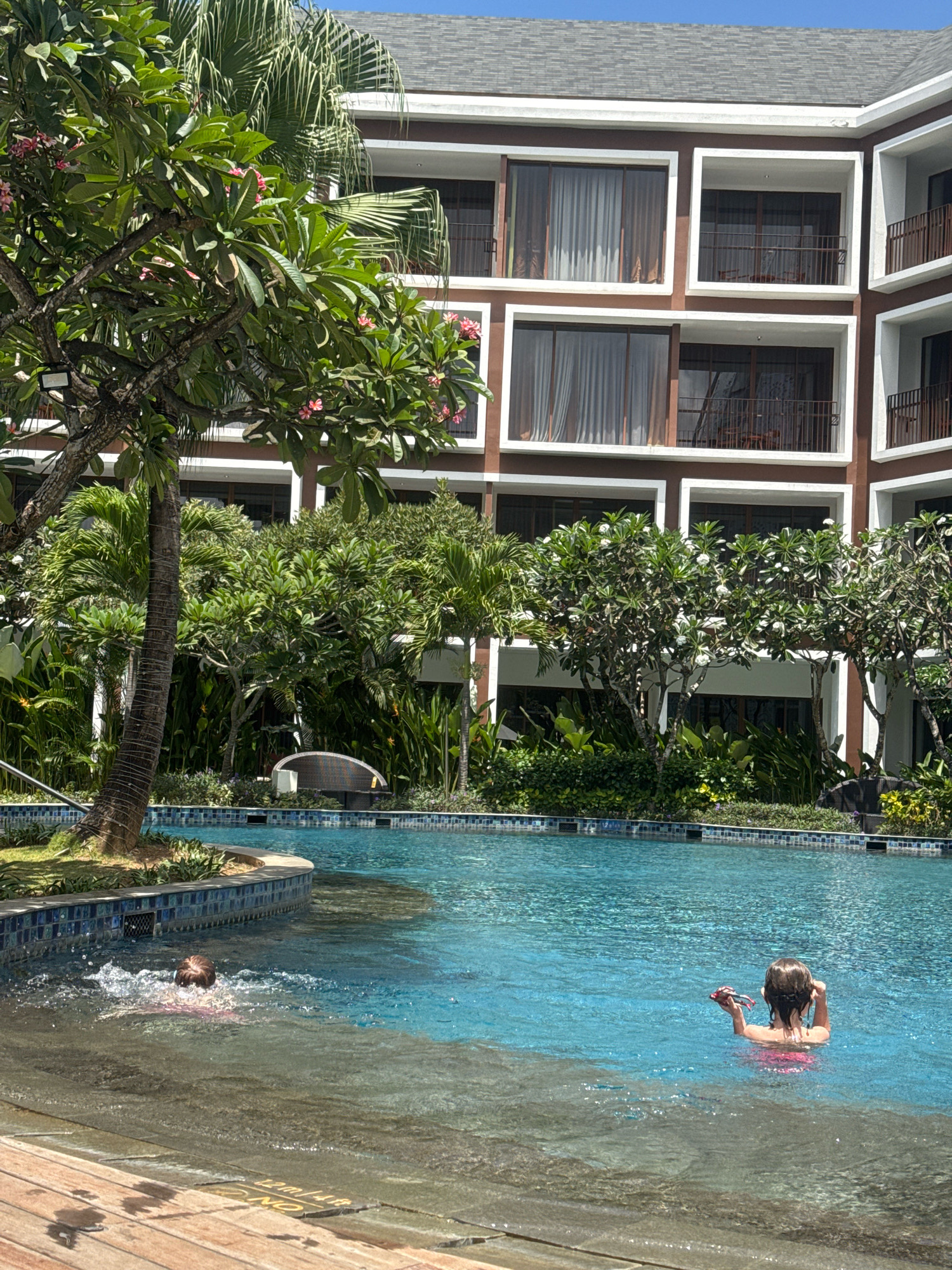 In Kuta, Indonesia, a resort swimming pool is captured on a sunny day. The clear blue water is partially in shadow from the overhanging balconies of a multi-story brown building. Two children are in the pool: one, with light brown hair, is swimming with their head above water, creating ripples as they move towards the left of the frame. The second child, also with light brown hair, is facing away from the camera, looking towards the right side of the pool, and holding a small, dark object. Lush green trees and plants surround the pool area, with a palm tree visible on the left. The pool is bordered by a tiled edge, and a wooden deck is present in the foreground.