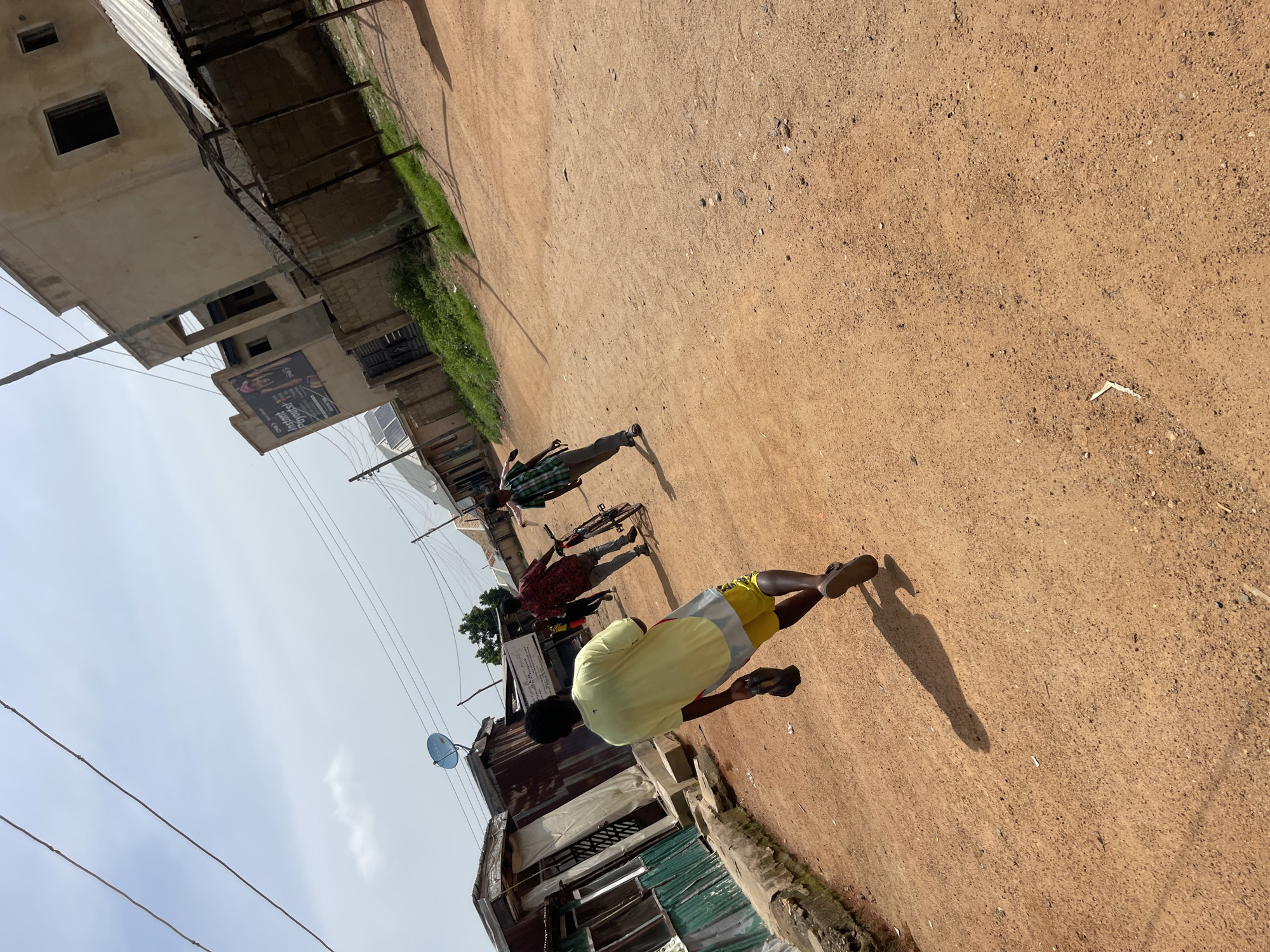 The image depicts a sunlit scene on a dirt road in Jos, Nigeria. Three young men are visible. In the foreground, a person in a yellow t-shirt and yellow and grey shorts is walking away from the camera, casting a long shadow on the dusty ground. To their left, further down the road, two other individuals are present. One is wearing a checkered shirt and appears to be standing or walking, with a bicycle nearby. Another person is seen in a dark, possibly red, patterned shirt, also on the road.
The environment suggests a casual, possibly residential or commercial area. Buildings with corrugated metal roofs and concrete structures line one side of the road. Some of these buildings appear to be shops or homes, with visible windows and signs. One sign has some visible text, appearing to be an advertisement with images of people and English text that is difficult to fully decipher but seems to include "INSTANT" and "JOYFUL". Electrical wires are strung overhead, typical of urban or semi-urban settings.
The sky is overcast with some patches of blue, suggesting it is daytime, likely late morning or early afternoon, given the strong shadows. The ground is dry and dusty, indicating warm, possibly dry weather. The overall atmosphere appears relaxed and everyday.
