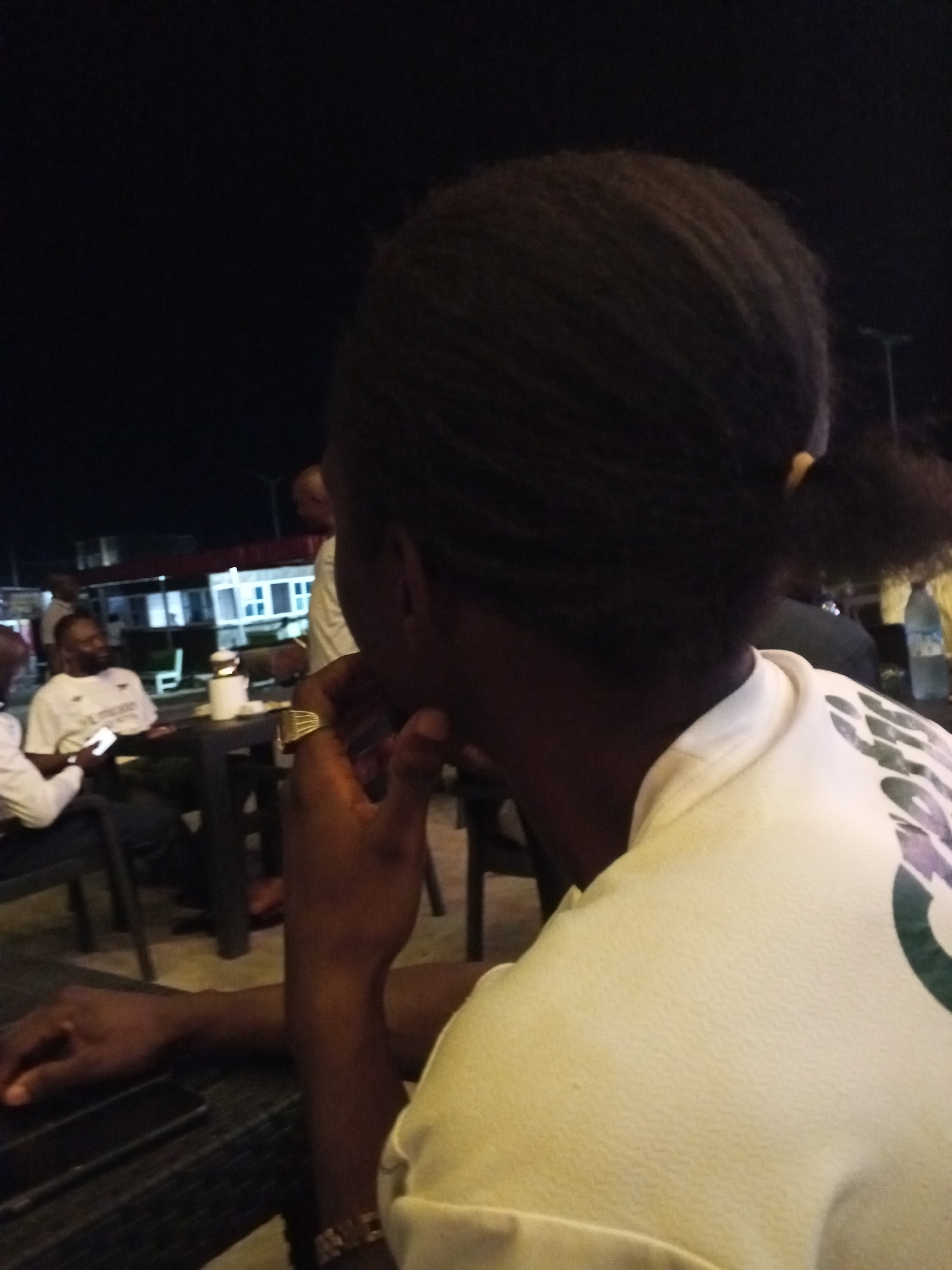 The photo shows the back of a person seated at a table outdoors at night, likely at a restaurant or bar. The person has dark hair pulled back into a ponytail and is wearing a white shirt with a green and purple design on the back. Their hand is near their face, and they are wearing a gold ring and a watch. A black smartphone is on the table. In the background, other people are seated at tables, and there is a building with bright lights visible. The location is possibly Lokoja, Nigeria. Details about interactions or specific actions of the individuals are not evident.