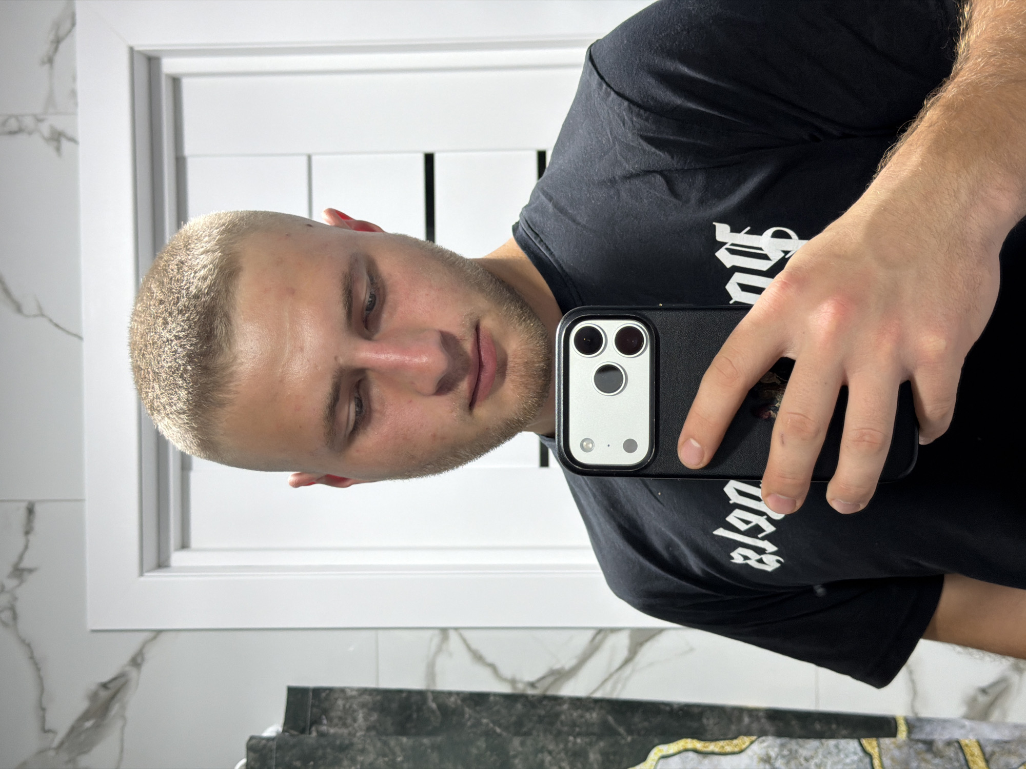A young man with blonde hair and a short buzz cut takes a selfie in a bathroom. He is wearing a black t-shirt with white lettering. He is holding a smartphone in his right hand, angled to capture his reflection. The background shows a tiled wall and a white doorframe. The lighting suggests an indoor setting, likely a home bathroom. The image is taken in Strășeni, Moldova.