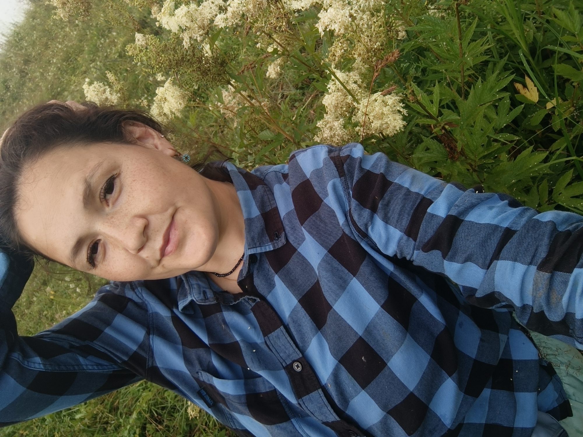 A young woman with brown hair and freckles lies on her back in a grassy field, with her head tilted towards the camera. She is wearing a blue and black checkered shirt and a beaded necklace. Her left arm is bent at the elbow and resting behind her head, while her right arm is bent and also behind her head. She is smiling and looking directly at the camera. In the background, there are white flowers and green foliage. The image appears to be taken outdoors, likely in a park or natural setting.