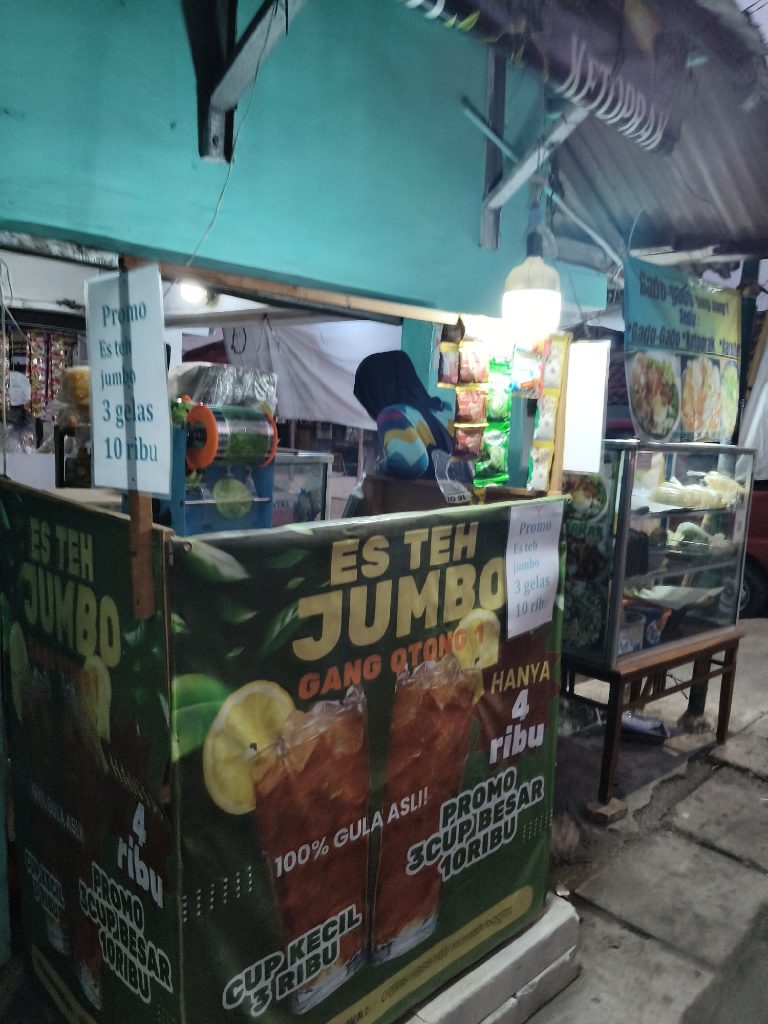 This is an outdoor, nighttime, medium shot of a street food stall in South Tangerang, Indonesia. The stall is illuminated by a bright, hanging lamp and the ambient light from the surrounding area. The stall features a green tarp with large text advertising "ES TEH JUMBO" (Jumbo Iced Tea) and "GANG OTONG" (Otong Alley). There are prominent graphics of iced tea glasses with lemon slices, and promotional text such as "HANYA 4 ribu" (Only 4 thousand Rupiah) and "PROMO 3 CUP BESAR 10 RIBU" (Promo 3 Large Cups 10 Thousand Rupiah).
A white sign board is propped up on a wooden post, displaying "Promo", "Es teh jumbo", "3 gelas 10 ribu" (3 glasses 10 thousand Rupiah). To the right of the main stall, a glass display case contains food items. Behind the stall, shelves are stocked with various packaged snacks and drinks. A person wearing a dark shirt is partially visible behind the stall, facing away from the camera. The ground in front of the stall is paved with concrete.