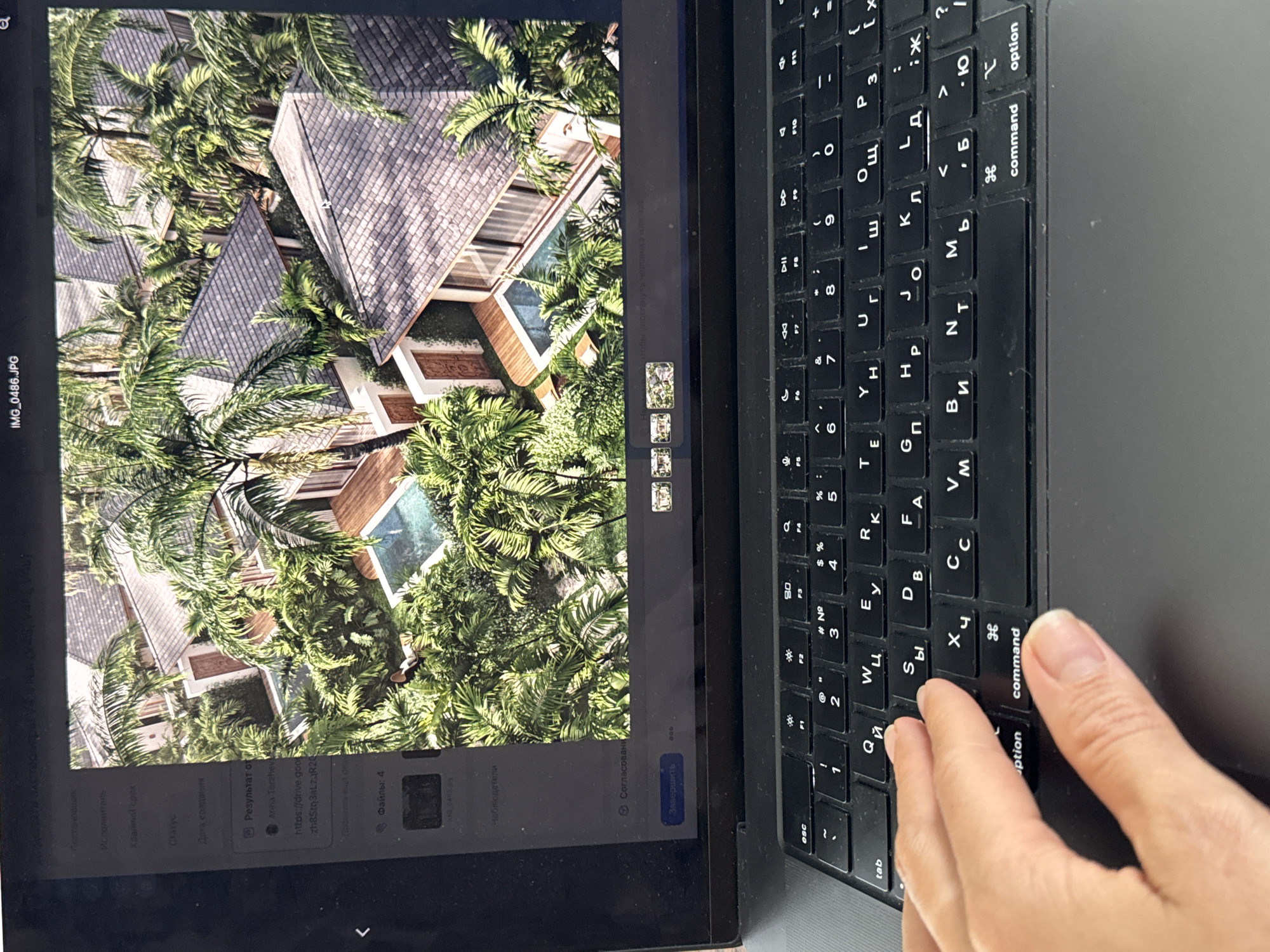 A photograph of a laptop and a tablet, positioned side by side on a gray surface. The tablet screen displays an aerial view of a property amidst dense tropical vegetation, possibly located in Denpasar, Indonesia. The scene features buildings with dark-tiled roofs partially obscured by palm trees and other greenery. A right hand is partially visible, resting on the keyboard of the laptop, which features keys labeled in both English and Russian. The laptop screen is dark.