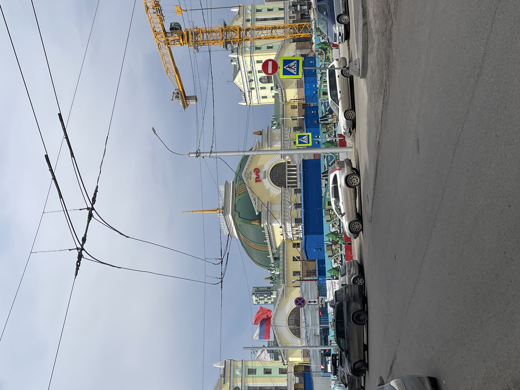 The image shows the Krasnoyarsk Railway Station, Russia. The station's facade features a green dome and yellow and white detailing. In front of the station is a street with several parked cars. Street signs are visible, including pedestrian crossing signs and a no-entry sign. A yellow construction crane is positioned near the building. Above the street, there are overhead power lines. The sky is overcast. The Russian flag is visible.