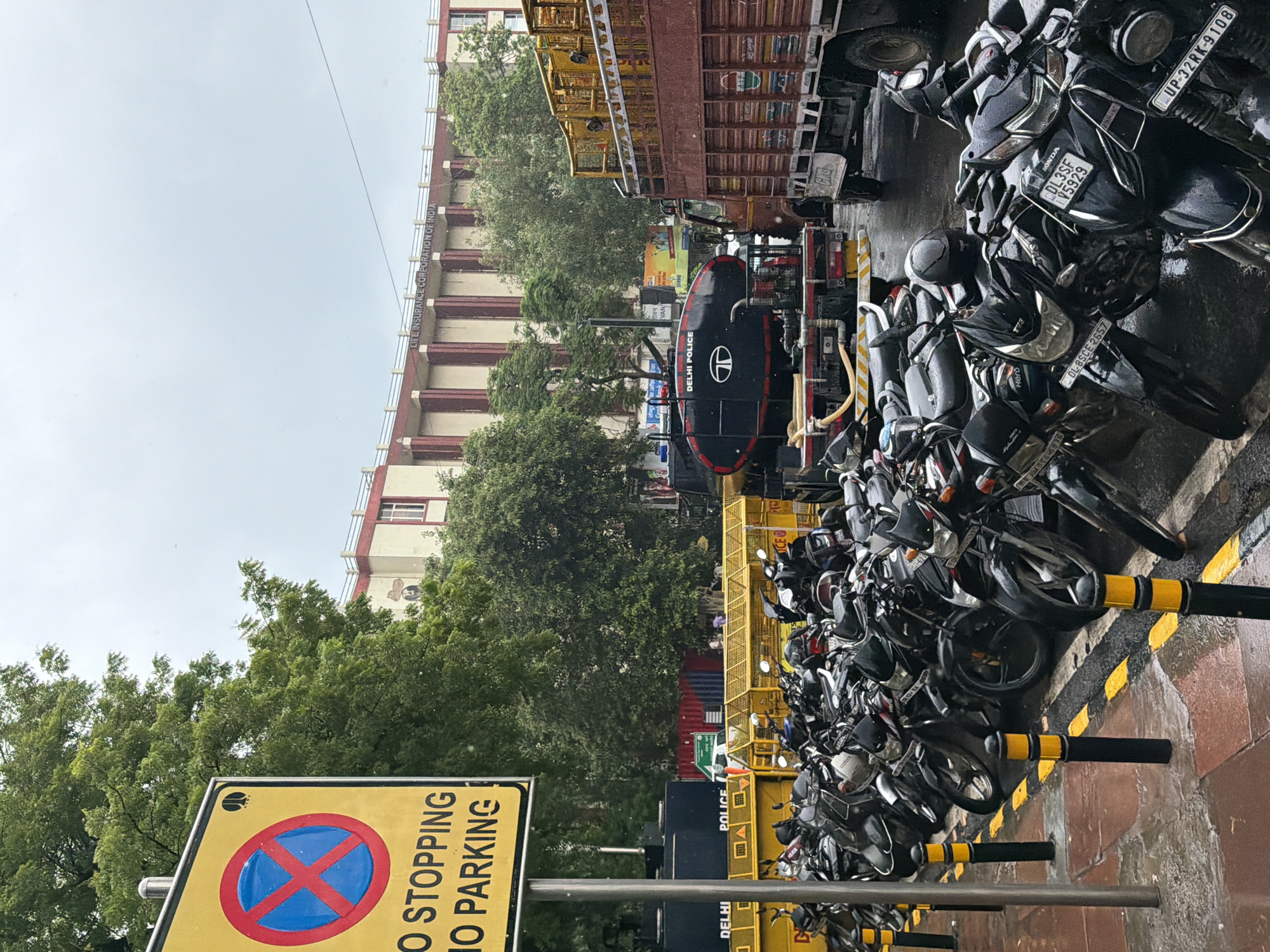 A street in New Delhi, India, is depicted on an overcast day, with a focus on a row of parked motorcycles. A yellow sign with a red circle and a blue "X" symbol, along with the text "NO STOPPING NO PARKING," is prominently displayed in the foreground on the left. Behind the motorcycles, a yellow police barricade stretches across the scene. Several dark-colored trucks are also visible, including one with a large black tank labeled "DELHI POLICE" and a Tata logo. In the background, a large, multi-story building with columns and red trim is present. The ground is wet, suggesting recent rain. A few individuals are standing or walking in the distance. The image captures a typical urban scene with traffic control measures and vehicles.