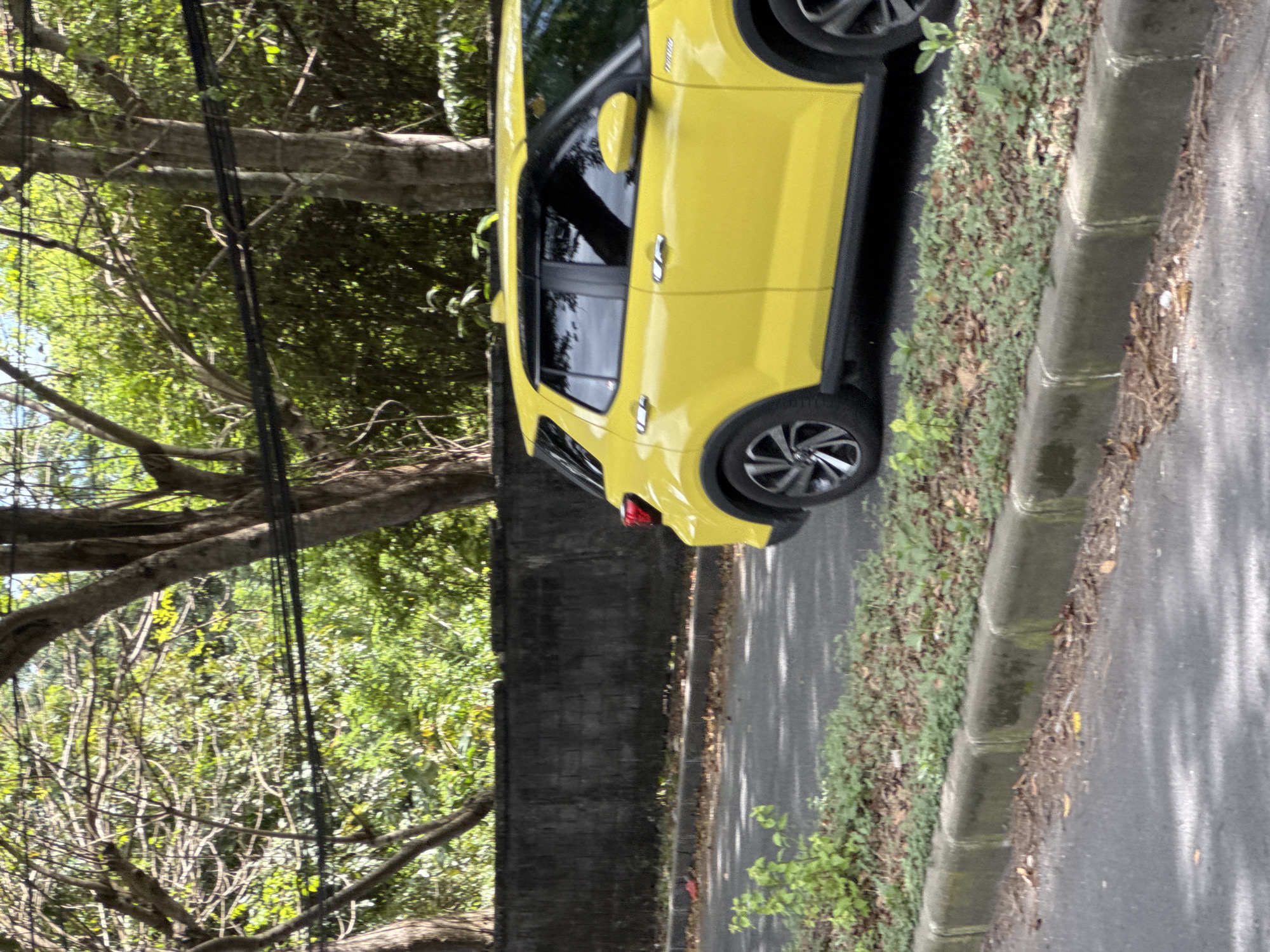 The image shows a yellow car parked on the side of a road next to a curb and some vegetation. A wall runs alongside the road, and beyond the wall, there are trees. Utility lines run through the trees. The location appears to be an urban or suburban area in a place called "Unknown, Unknown".