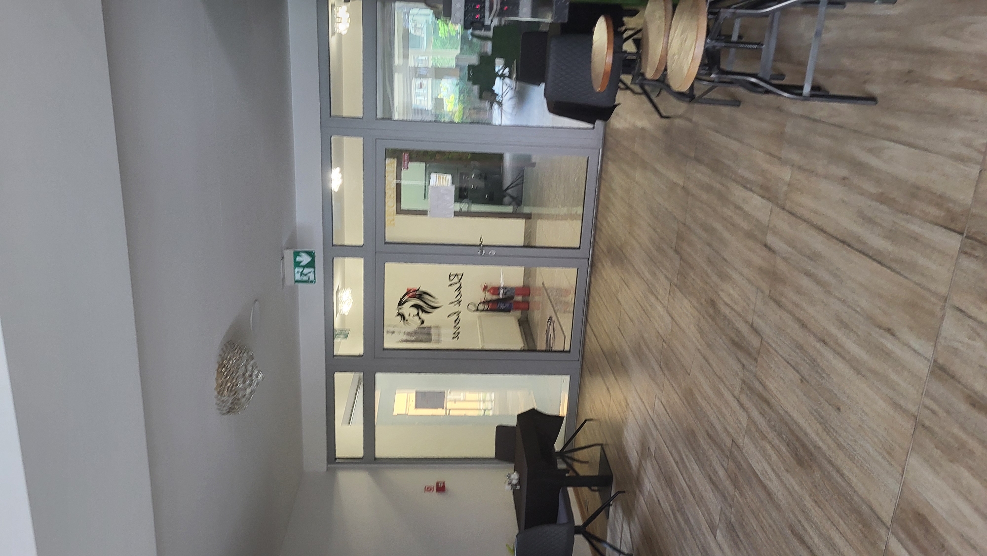 The image shows an interior hallway or lobby area of a building with a glass-paneled entrance. The floor is covered in light brown wood-look tiles. To the left, there is a small square table covered with a black tablecloth and two dark chairs. Further left on the wall is a red fire alarm pull station.
The central feature is a set of double glass doors with gray frames. On the left door, a black logo of a horse's head with a red mane is visible, along with the text "Black Lion" in mirrored script. Two red fire extinguishers are standing in front of the left door. The right door leads to an area labeled "RECEPCIA" (Reception in Slovak), with a counter and chairs visible in the background. There are also two green exit signs with arrows pointing downwards above the glass doors.
To the right of the entrance, there are three bar stools with dark metal frames and light wooden seats. The background through the glass doors reveals an outdoor area with seating and greenery. The ceiling is white, and a decorative chandelier hangs near the left side of the image. The overall impression is a modern, clean interior. This location is likely the reception area or waiting room of a business, possibly a salon or restaurant, in Prešov, Slovakia.