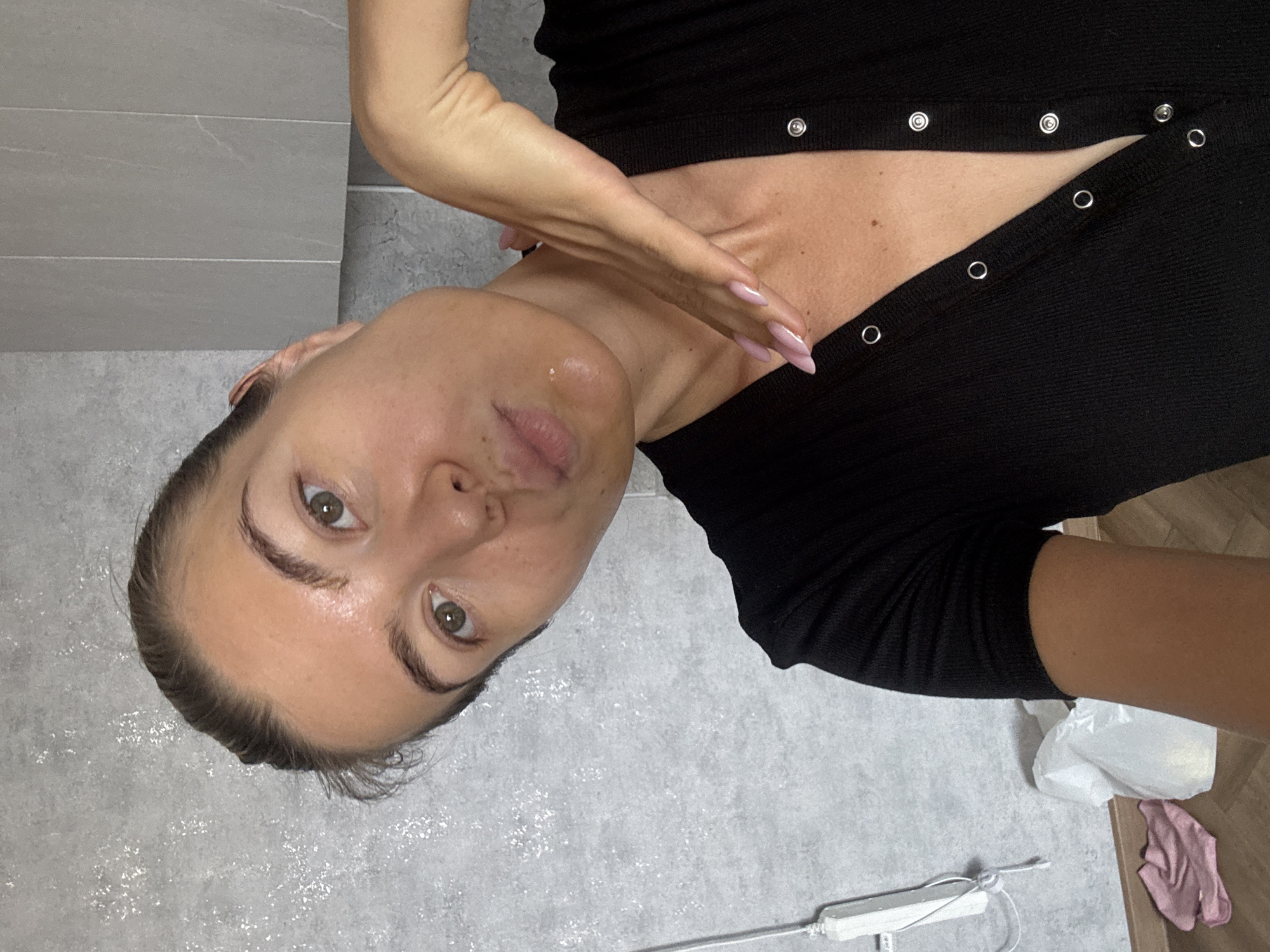 A young woman with her hair pulled back is taking a selfie in a dimly lit room. She is wearing a black, ribbed, short-sleeved top with silver snaps down the front. Her left hand is resting on her neck, and her right hand is raised to her face. The background is a gray tiled wall. To the left of the woman, a white plastic bag and a pink cloth are on the floor, and a white electrical cord is attached to the wall.