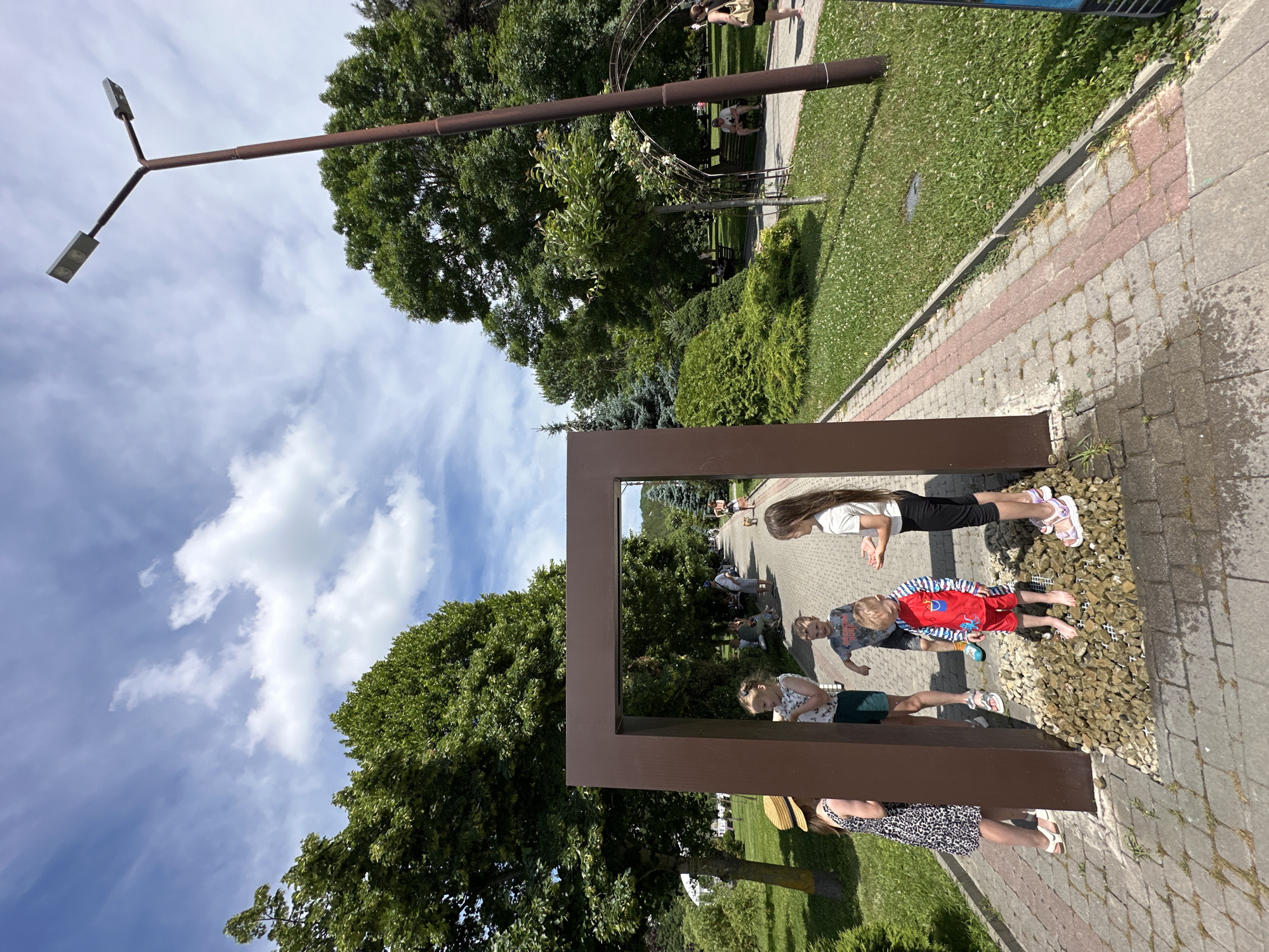 The image depicts a sunny day in a park, likely in Abrau-Dyurso, Russia, given the context. A large, brown, rectangular frame is the central focal point, positioned on a walkway paved with interlocking stones and bordered by a strip of pinkish stones. The frame stands on a bed of pebbles, through which water appears to be flowing from a grate.
Several children are gathered around this frame, interacting with it and each other. On the left, a young girl wearing a straw hat and a black and white patterned dress stands with her back to the camera. To her right, inside the frame, a girl in a white sleeveless top and dark shorts is visible, looking towards the right. In front of them, a boy in a red and blue striped swimsuit stands barefoot on the pebbles, looking down with a slightly distressed expression. To the right of the boy, a girl with long dark hair, wearing a white t-shirt and black leggings, looks towards the boy. Another boy, wearing a grey t-shirt with a graphic print, is partially visible behind the girl in the white top, appearing to gesture.
The park setting features lush green trees and manicured lawns with small white flowers. A paved path, similar to the one in the foreground, runs alongside the grassy area. In the background, people are seen relaxing on benches and walking. A tall, dark brown lamppost with two lights is prominently positioned to the right of the frame, casting a shadow on the path. An arched trellised structure covered in greenery is also visible in the background. The sky is blue with scattered white clouds.