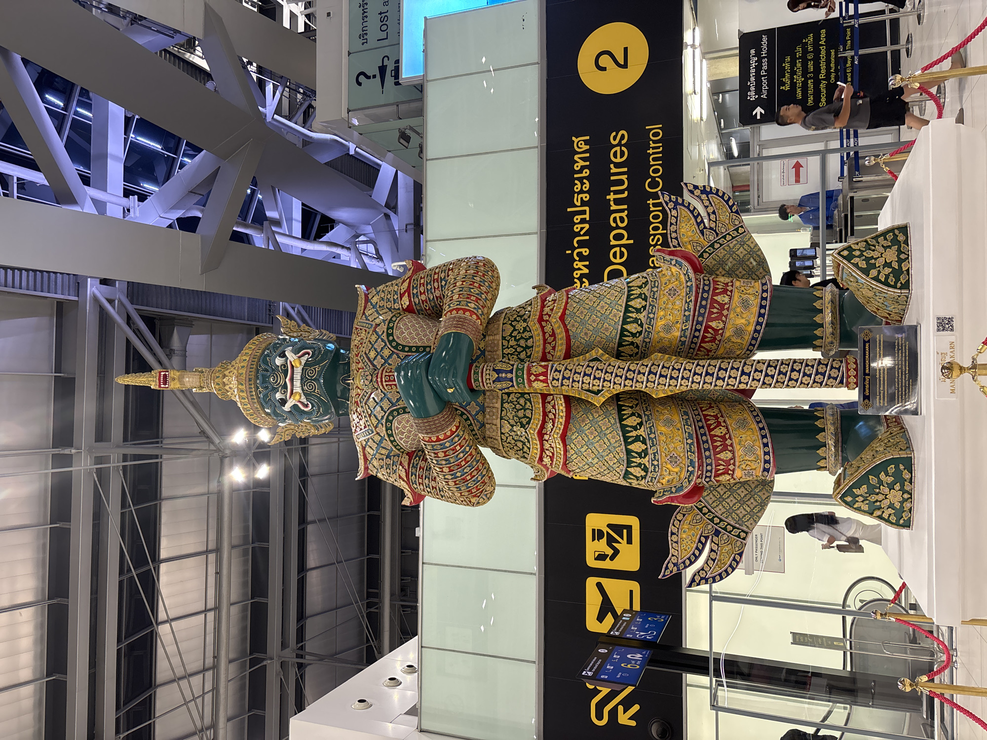 The image captures a large, ornate statue of a green-skinned giant, likely a yaksha from Thai folklore, standing in what appears to be an airport terminal. The statue is richly decorated with intricate patterns in gold, red, and blue, and it holds a large weapon. In the background, there are airport signs indicating "Departures" and "Passport Control," along with directional signs and a large number "2" in a yellow circle. The architecture of the terminal is visible, with high ceilings, exposed beams, and large windows. Several people are present in the scene, including travelers walking by, airport staff at security checkpoints, and individuals interacting with information displays. The lighting suggests an indoor environment. The location is identified as Nong Prue Subdistrict, Thailand, which is part of the Bangkok Metropolitan Region, a common area for major international airports.