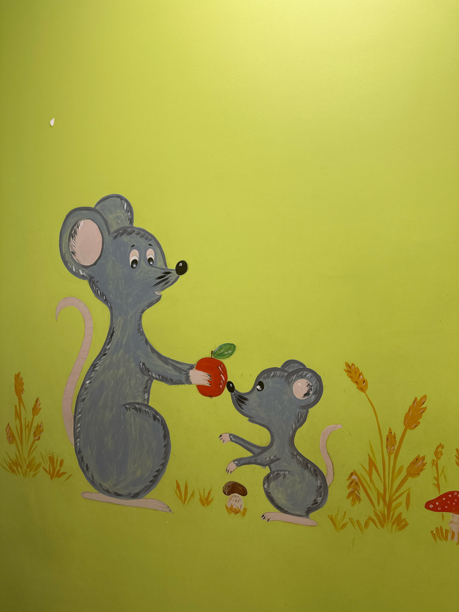 A mural painted on a pale green wall depicts two cartoon mice in a stylized outdoor scene. A larger gray mouse on the left is offering a red apple to a smaller gray mouse on the right. The larger mouse holds the apple in its right hand, and its left paw is outstretched. The smaller mouse is reaching out with its left paw towards the apple. Both mice have pink inner ears, black eyes, and black noses. The larger mouse has a long, pink tail that curves upwards. The smaller mouse has a similar, shorter tail.
The background of the mural includes elements of nature painted in yellow and brown. To the left of the larger mouse, there are strokes of yellow paint that resemble grass. To the right of the smaller mouse, there are stalks of wheat or grain, also painted in yellow and brown. Two mushrooms are depicted on the ground: a brown-capped mushroom to the left of the smaller mouse and a red-capped mushroom with white spots to the far right. The wall itself is a pale, slightly textured green. There is a small, bright white mark on the upper left quadrant of the wall, appearing to be a smudge or imperfection on the paint. The scene is consistent with a children's mural and is likely located in Vidnoye, Russia.