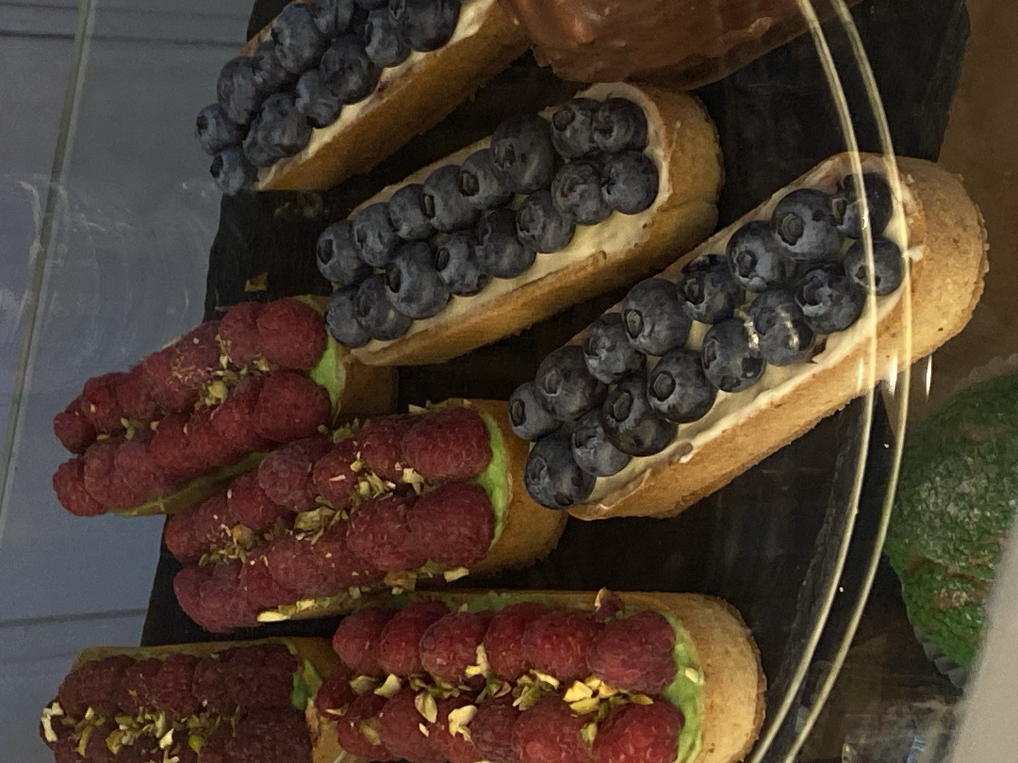 A close-up, slightly angled shot showcases a display of assorted pastries in a refrigerated case. The pastries are rectangular in shape and appear to be tartlets. Several are topped with vibrant red raspberries and chopped pistachios, set against a layer of green cream. Others are adorned with plump blueberries, also on a cream base. The pastries are arranged on a dark surface, with a reflective clear barrier in the foreground. The background is blurred, showing a hint of vertical lines, possibly from the display case itself. No people are visible in the image. The scene appears to be a bakery or patisserie, likely in the city of Agronom, Russia, based on the provided context.