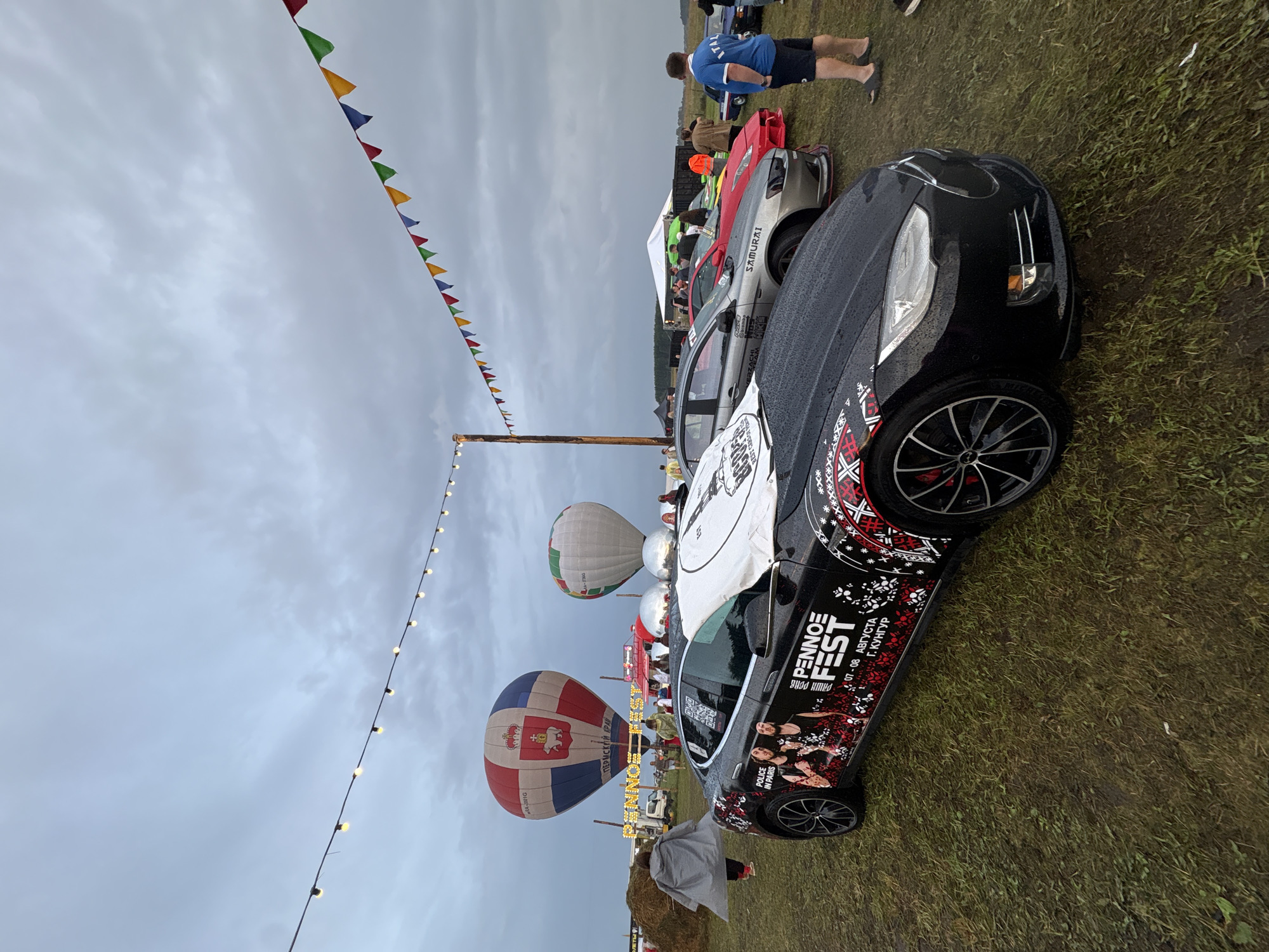 An outdoor event is taking place under a cloudy sky, with two hot air balloons visible in the background. The foreground is dominated by a black Tesla Model S, customized with "PENNOE FEST" graphics and a black and red ornamental design along the side. Other cars, including a silver Mitsubishi Lancer Evolution and a red sports car, are parked behind the Tesla. Several people are scattered throughout the scene, some appearing to be participants or attendees of the event. The ground is covered in grass, suggesting a rural or park-like setting. The overall atmosphere is one of a festive gathering.