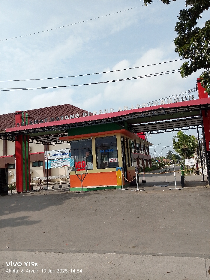 The image displays the entrance of a facility, identified as RSUD Arjawinangun (Regional Public Hospital Arjawinangun) in Indonesia, under a clear, bright sky with scattered clouds. The scene is illuminated by daylight, consistent with the timestamp of 2:54 PM on January 19, 2025.
A prominent entrance gate spans across the road, featuring a red and green archway. White lettering on the arch reads: "SELAMAT DATANG DI RSUD ARJAWINANGUN" (Welcome to Arjawinangun Regional Public Hospital). Beneath this arch, a colorful security booth painted in orange, green, yellow, and blue, is situated at the entry point. On its windows, several signs are visible, including "IGD" (Emergency Room), "POS SATPAM" (Security Post), and "PELAYANAN KUNCI" (Key Service or a service counter).
To the left of the booth, part of a multi-story building, likely the main hospital, can be seen. It features a brown tiled roof and what appears to be a medical cross symbol on its side. Banners with largely unreadable text are affixed to this building. The foreground shows an asphalt road leading into the complex. White traffic barriers, linked by yellow chains, are placed on the road, indicating controlled access. Beyond the gate, the road extends further into the facility, flanked by other structures and some trees. No specific human activity is clearly visible, suggesting a moment without active entry or exit. The overall impression is of a functional public health institution.