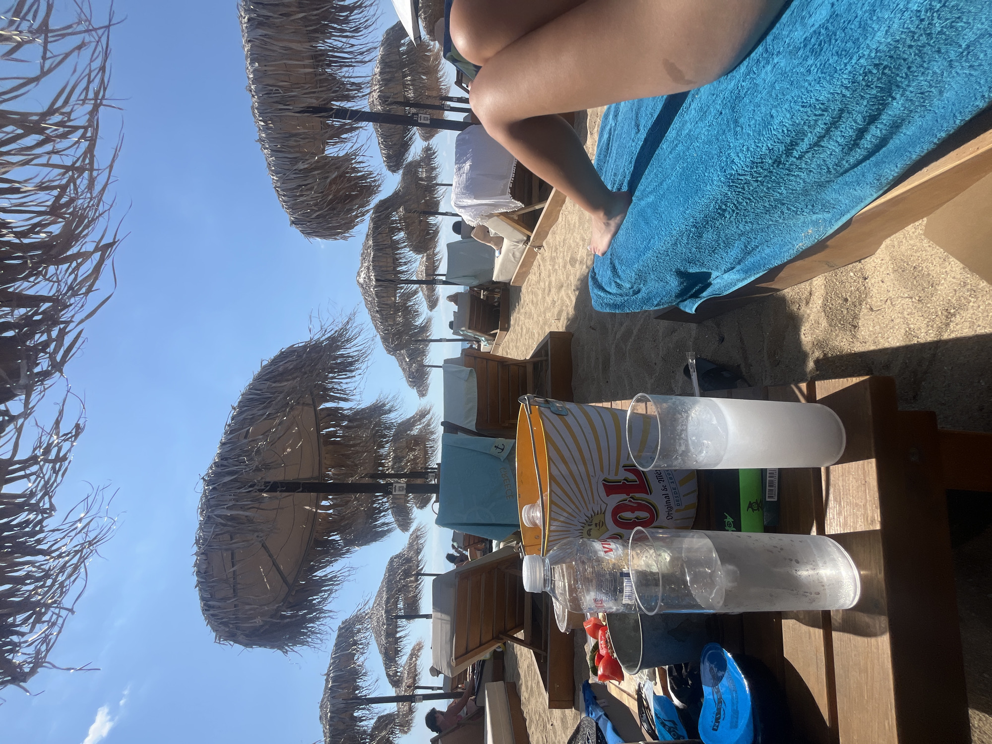 The image depicts a beach scene in Νέα Ηράκλεια, Greece, on a sunny day. In the foreground, a wooden table holds two glasses of clear liquid with ice, a bottle of water, a bucket with the "Sol" logo, and some halved tomatoes. A person's legs, covered by a blue towel, are visible on the right, suggesting they are lounging on a beach chair. In the background, rows of thatched umbrellas shade numerous empty beach chairs, with a few people scattered further down the beach. The sandy ground and clear blue sky complete the tranquil, seaside atmosphere.