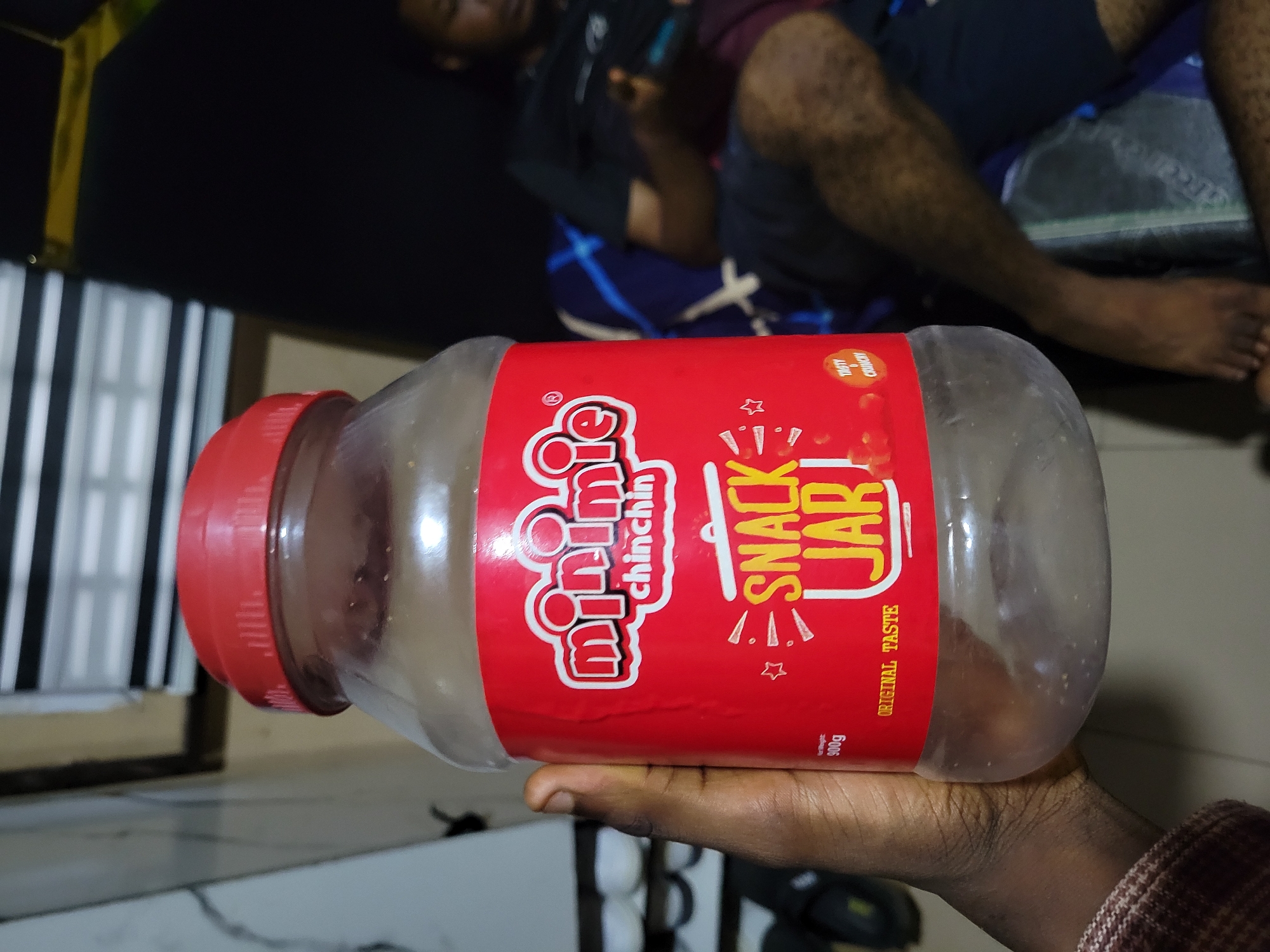 The image shows a close-up of a large, clear plastic jar with a red lid and a prominent red label. The jar is held by a person's hand, which is positioned in the lower right corner of the frame. The jar's label is mostly visible and features bold white and yellow text. The largest text reads "SNACK JAR" in a playful font, with "minnie" in smaller white letters above it. Below "SNACK JAR" and to the right, the words "ORIGINAL TASTE" are printed vertically. There are also smaller words like "chinchin" and some decorative stars and lines on the label. The jar appears to be nearly empty, with some residue visible on the inside.
In the blurred background, two individuals are partially visible, reclining on a dark surface, possibly a bed or sofa. One person is lying down with their legs extended, wearing dark shorts with a blue pattern. The other person is partially visible behind them, also appearing to be resting. The lighting suggests it might be evening or indoors, with artificial light casting shadows. The overall scene has a casual, domestic feel, and the focus is clearly on the snack jar. No specific weather conditions or strong emotional cues are evident, other than the implied satisfaction of enjoying snacks. The visible text is primarily focused on the product branding and description.