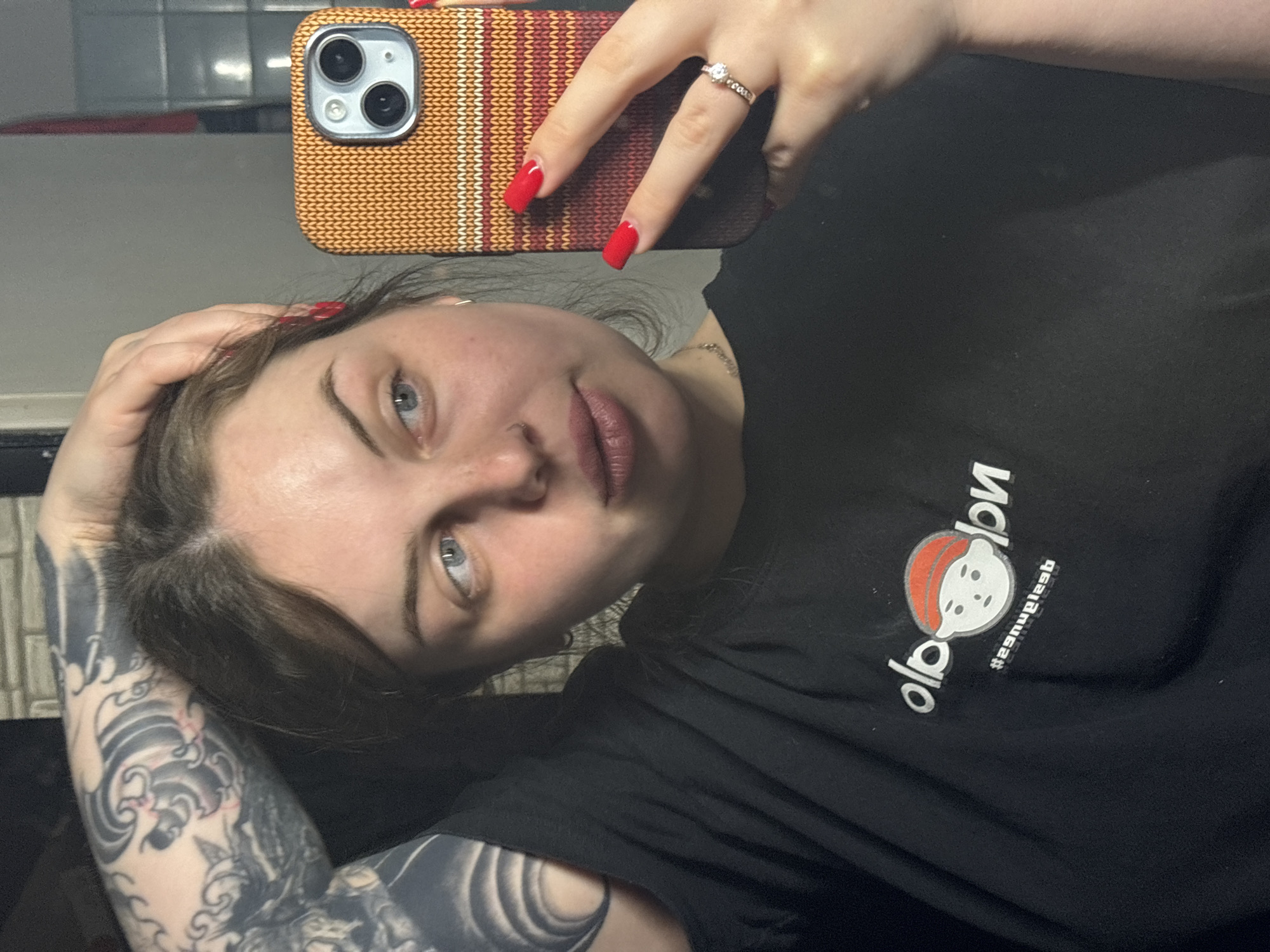 A fair-skinned woman with blue eyes and dark hair is taking a selfie with a phone in a knit-textured orange and brown case. She wears a black T-shirt with a white, cartoon-style graphic and text, and a ring on her finger. Tattoos cover her left arm. The background is partially blurred, showing a tiled wall. The location cannot be determined from the image.