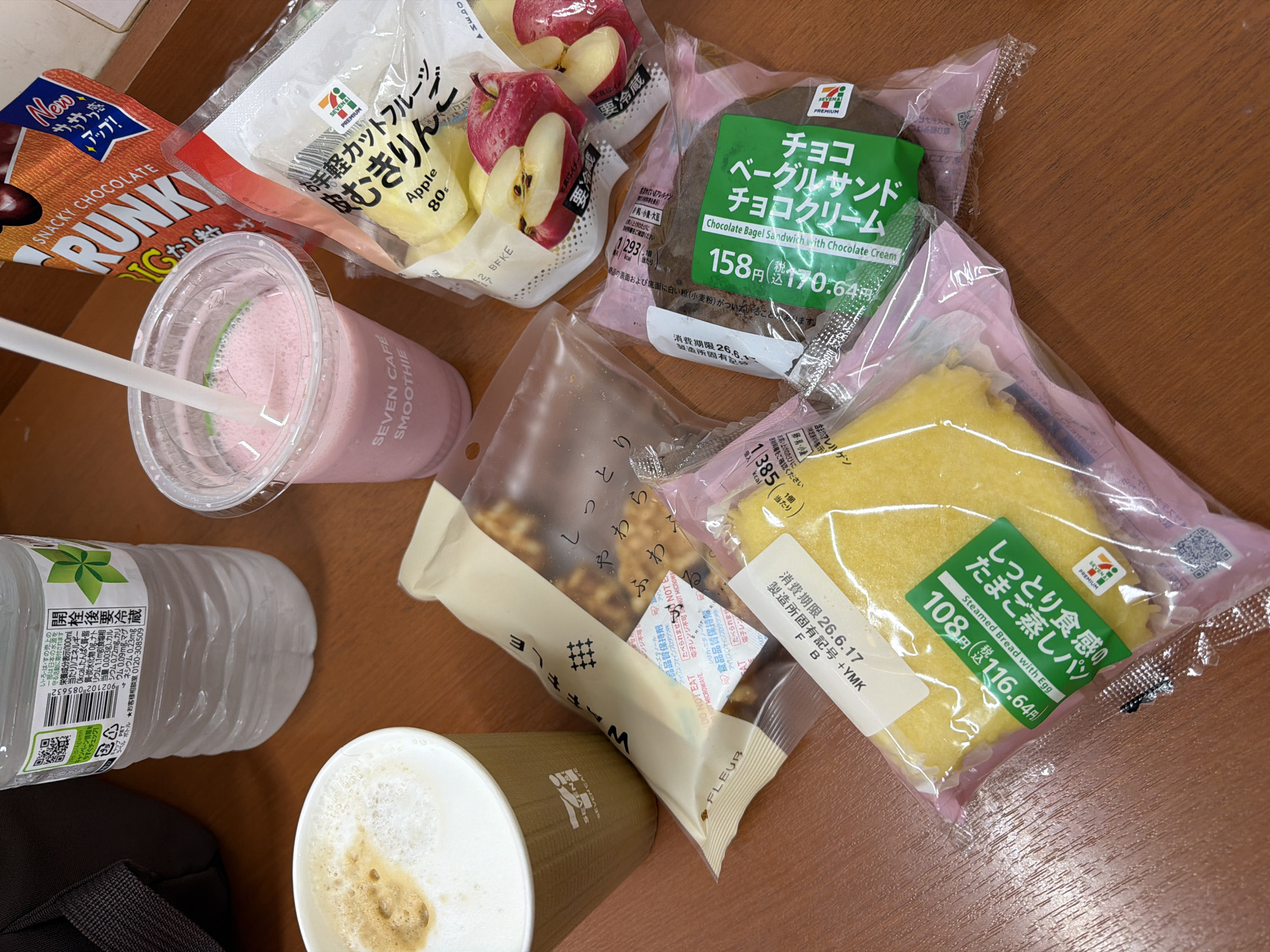 The image displays a selection of convenience store food items laid out on a wooden surface, likely a table. In the upper left corner, a portion of a black bag is visible. To its right, a clear plastic bottle of water with a white label containing Japanese text and a QR code is present. In the foreground, a white paper cup with the "SEVEN CAFE" logo holds a frothy, light brown beverage, possibly coffee or latte. Next to it, a clear plastic cup with a white lid and straw contains a pink smoothie, also with the "SEVEN CAFE SMOOTHIE" branding.
Several packaged food items are arranged around these drinks. To the right of the coffee cup, a beige and white striped wrapper encloses a long, narrow pastry. Further right, a bag of sliced apples is visible, with two whole apples and several apple slices displayed. Above the apples, an orange and white package labeled "SNACKY CHOCOLATE" is prominent.
Below the apples and smoothie, a dark brown, round pastry in a clear plastic wrapper is labeled "チョコレートクリーム" (Chocolate Cream) and "ベーグルサンド" (Bagel Sandwich). To the left of this, a yellow, rectangular cake-like item in a pink and clear wrapper is visible, with a green label that reads "たまご蒸しパン" (Steamed Bread with Egg). A white label with Japanese text, including a date and characters like "+YMK", is attached to this yellow pastry.
The overall scene suggests a casual meal or snack break, possibly at a convenience store or a seating area within one. The lighting is natural and even, and the image is taken from a slightly elevated, top-down perspective. The context of "Chuo, Japan" for the city location indicates a potential real-world setting where these items might be purchased or consumed.