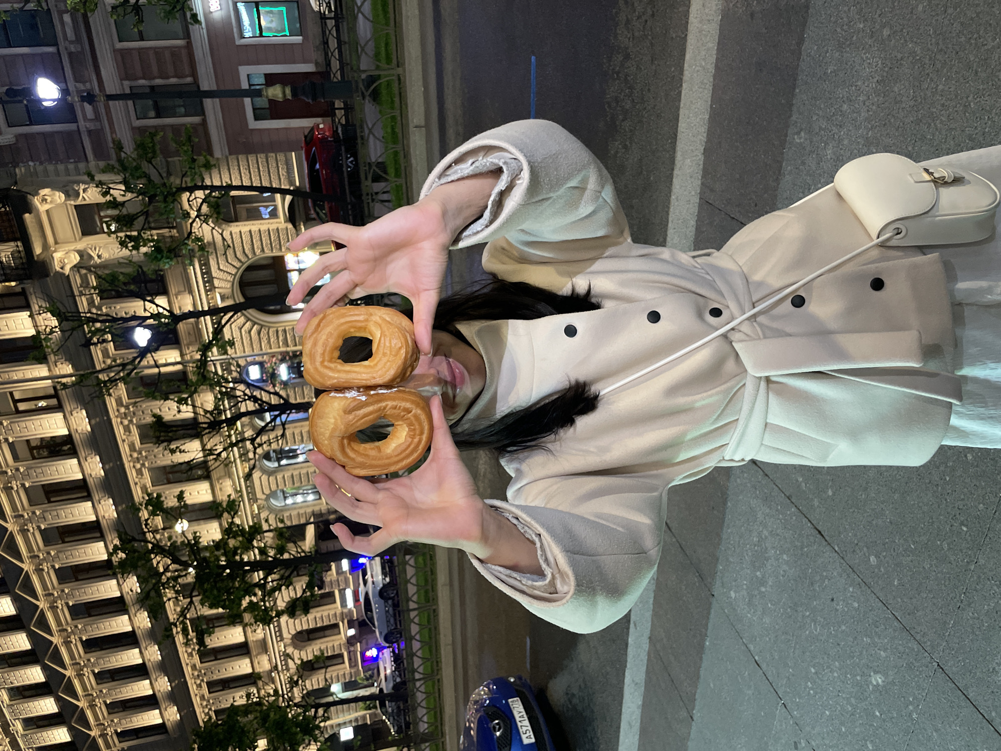 In an urban setting at night, a young woman stands on a sidewalk, holding two ring-shaped pastries up to her eyes. She is wearing a cream-colored trench coat cinched at the waist with a matching belt, and a small white crossbody bag. In the background, a multi-story building with illuminated windows and ornate architectural details stands prominently. Streetlights cast a warm glow on the scene. The street in front of the building has vehicles, including a blue car with visible license plate "A571AY 776", and a red car further down. The architecture suggests a location in Saint Petersburg, Russia, likely in a historic district with classical building designs. The overall atmosphere is casual and playful, with the woman interacting with her surroundings.