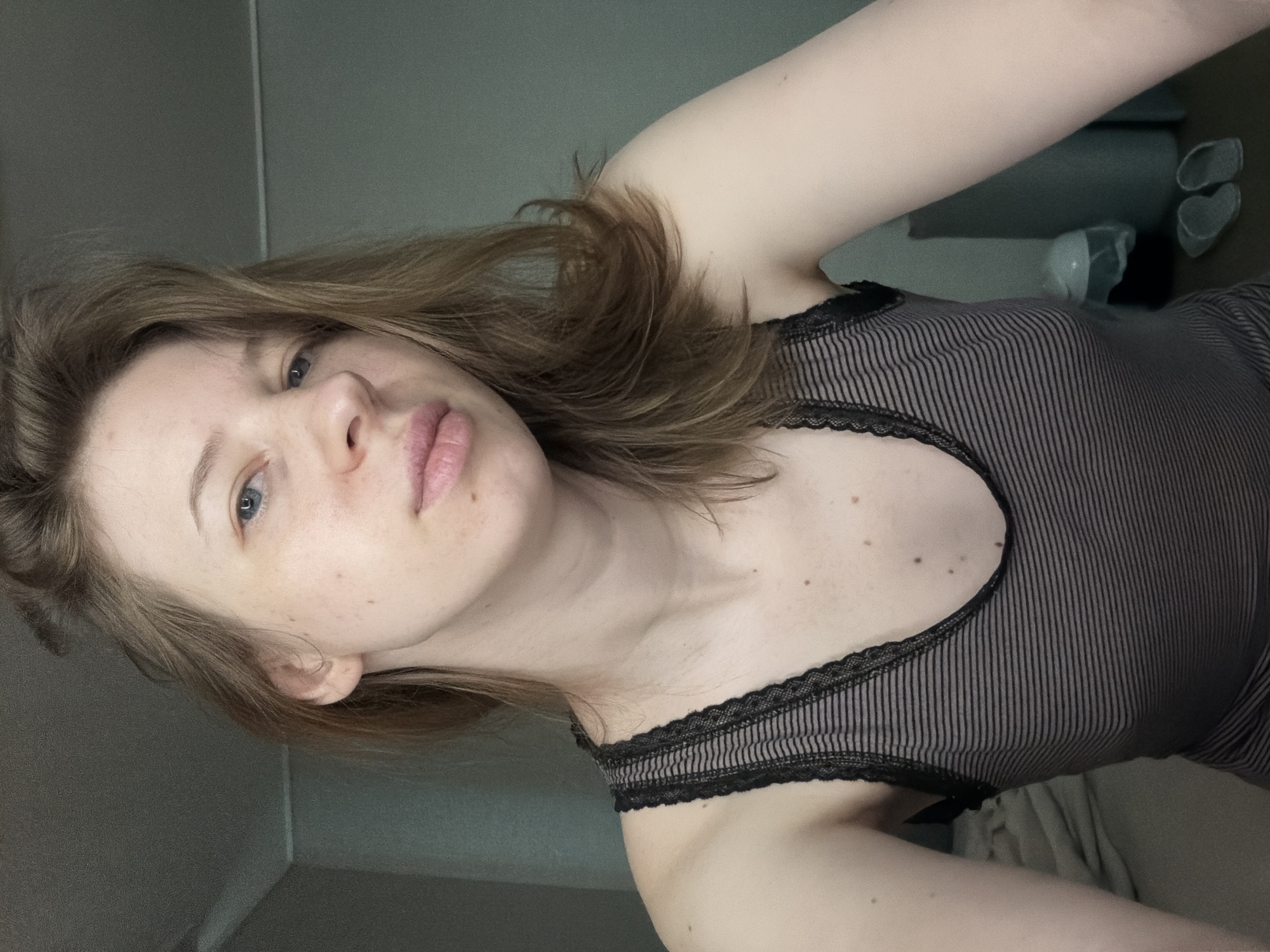 A young Caucasian woman with light brown hair, fair skin, and blue eyes is captured in a medium close-up shot. She is looking directly at the camera with a neutral expression. Her hair is styled loosely, falling around her shoulders. She is wearing a striped tank top with lace trim. The background appears to be an indoor setting, possibly a bedroom, with a plain wall and some bedding visible. The lighting is soft and natural, suggesting it might be daytime. The image was taken in Dongducheon-si, South Korea.