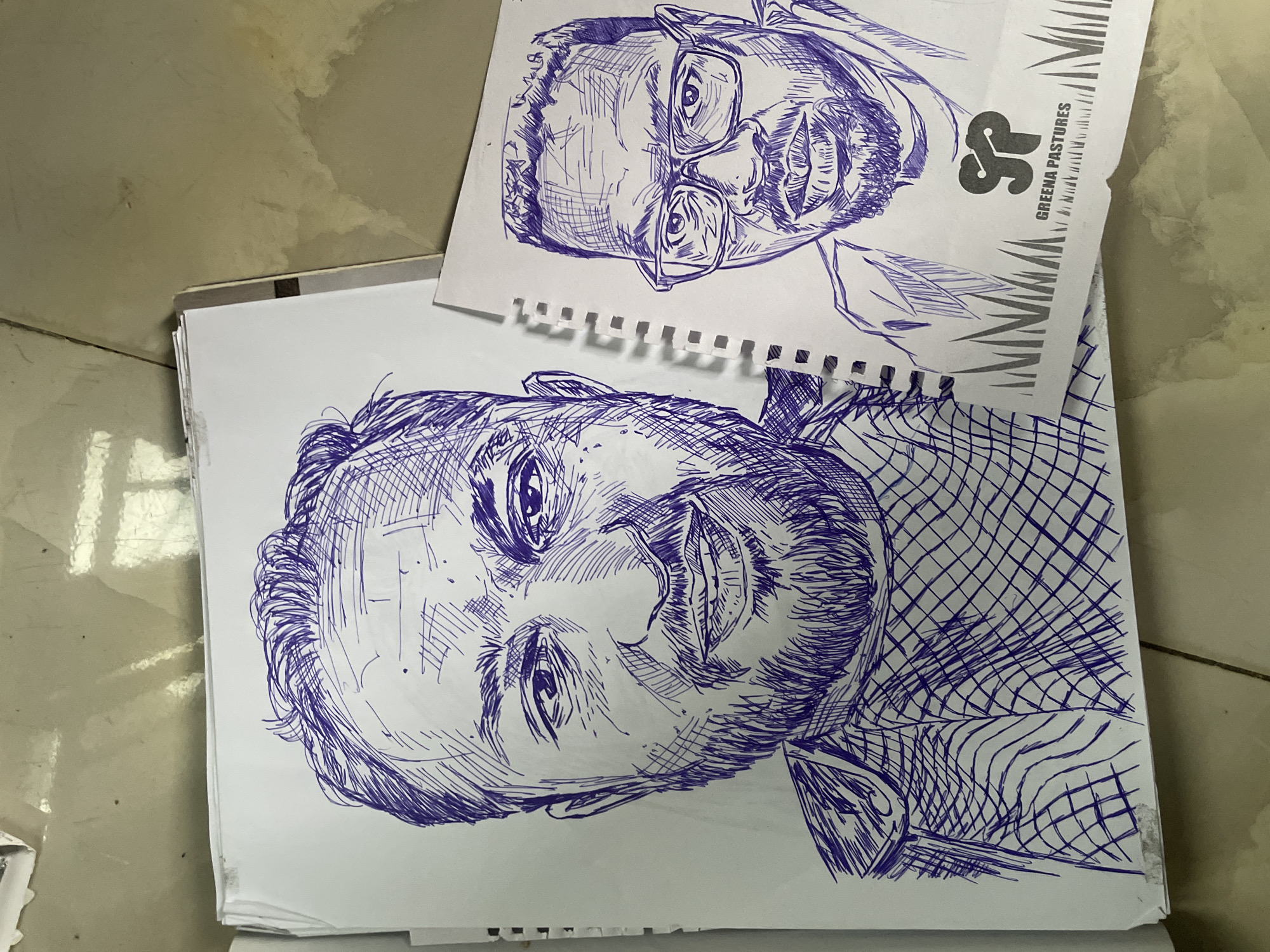 Two pen and ink portraits of men are depicted on white paper, with the paper laid out on a tiled surface. The portrait on the left is larger and faces the viewer directly, showing a man with a beard, mustache, and short hair. He is wearing a collared garment with a checkered pattern. The portrait on the right, smaller and on a separate sheet, also faces the viewer, this time a man wearing glasses, a beard, and a mustache. He has short, styled hair. Below his portrait, a logo with the text "SP GREENA PASTURES" is visible, set against a background of grass sketches. The tiling appears to be a light beige or cream color with visible grout lines, and the lighting suggests an indoor environment. The image captures the drawings from a slightly elevated, overhead perspective.