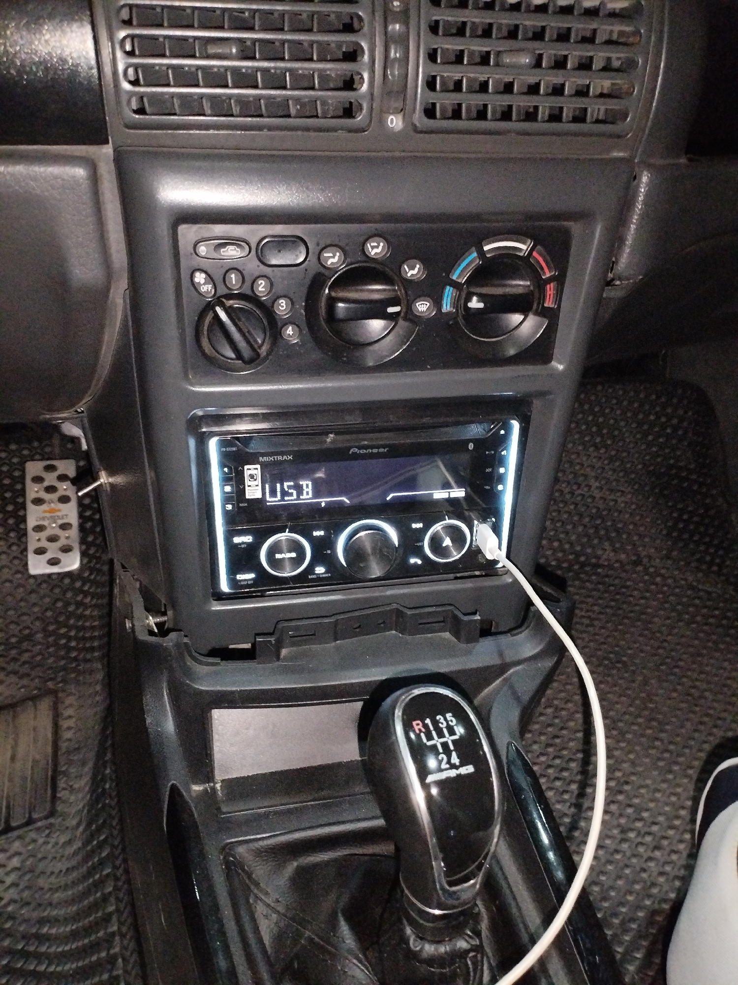 The image shows the interior of a car, specifically the dashboard area around the radio and gear shifter. A Pioneer car stereo with a USB port is prominently displayed, with a white USB cable connected to it. To the left of the stereo, there are several knobs and buttons controlling the car's climate system and a vent. Below the stereo, a gear shifter with markings for "R", "1", "3", "5", "2", and "4" is visible, indicating it is a manual transmission. A silver pedal with the Chevrolet logo is mounted on the floor to the left of the driver's seat. The floor mats are black and textured. The overall impression is of a functional, albeit somewhat aged, car interior. The scene does not provide any direct evidence to suggest it is located in Termez, Uzbekistan; this information is based on external context not present in the image itself.