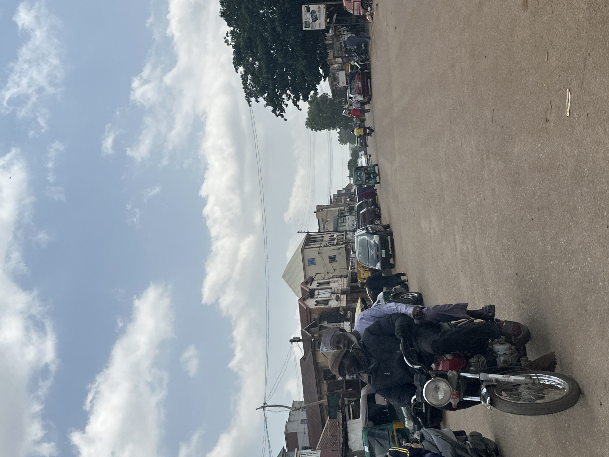 This image depicts a busy street scene in Jos, Nigeria. The foreground is dominated by a motorcycle carrying two people. The driver, wearing a dark jacket and a beanie, is focused ahead, while the passenger, dressed in a light purple shirt, is seated behind him. The motorcycle is red with silver accents and has a visible license plate.
The street is lined with buildings of varying architectural styles, suggesting a mix of older and newer constructions. Some buildings appear to be shops, indicated by signs and goods displayed. The road itself is paved and appears to be made of asphalt or a similar material.
Several other vehicles are visible, including more motorcycles, cars, and a three-wheeled auto-rickshaw. This indicates a bustling environment with active transportation. The presence of pedestrians walking along the street further contributes to the sense of a lively urban setting.
The sky is partly cloudy with patches of blue sky visible, suggesting it is daytime. The lighting indicates it could be late morning or early afternoon. The overall atmosphere appears to be one of everyday activity and commerce, with no overt signs of distress or celebration. The scene is typical of a busy commercial area in a Nigerian city.