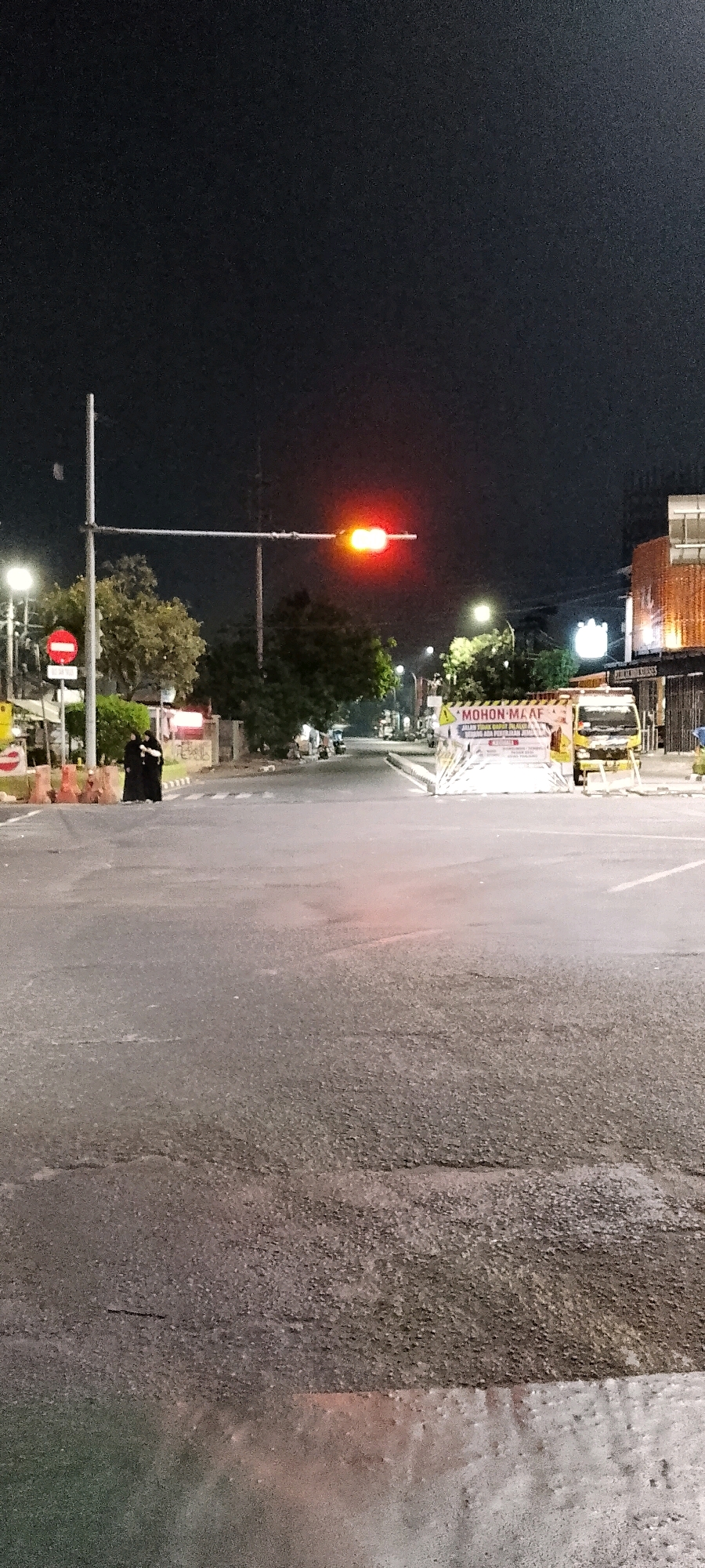 The image shows a street scene at night in Bekasi, Indonesia. A traffic light displays a red signal. Two individuals dressed in dark, traditional Islamic attire are standing on the left side of the road, near a traffic barrier. A yellow truck is parked on the right side of the road, behind a temporary barrier with a sign that reads "MOHON MAAF JALAN TIDAK DAPAT DILALUI ADA PERBAIKAN JALAN" (Sorry, the road cannot be passed, there is road repair). The street appears to be wet, reflecting the lights from the surroundings. Trees and buildings are visible in the background.