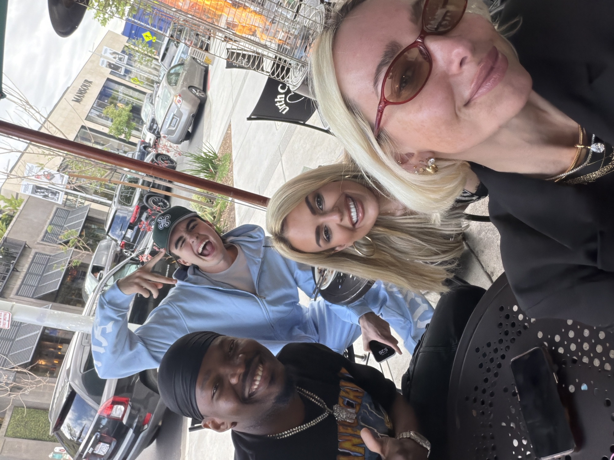 The image depicts a group of four individuals taking a selfie outdoors at an establishment in West Hollywood, California, United States of America. In the background, the "MANSOUR" storefront is visible. From left to right, a young man with a baseball cap and a blue sweatsuit is flashing a hand gesture. Below him, another man with a black durag and a graphic T-shirt is smiling. Next, a blonde woman is seen smiling into the camera, and the person taking the photo is a woman with blonde hair and red glasses. A phone lies on the table in the bottom right corner.