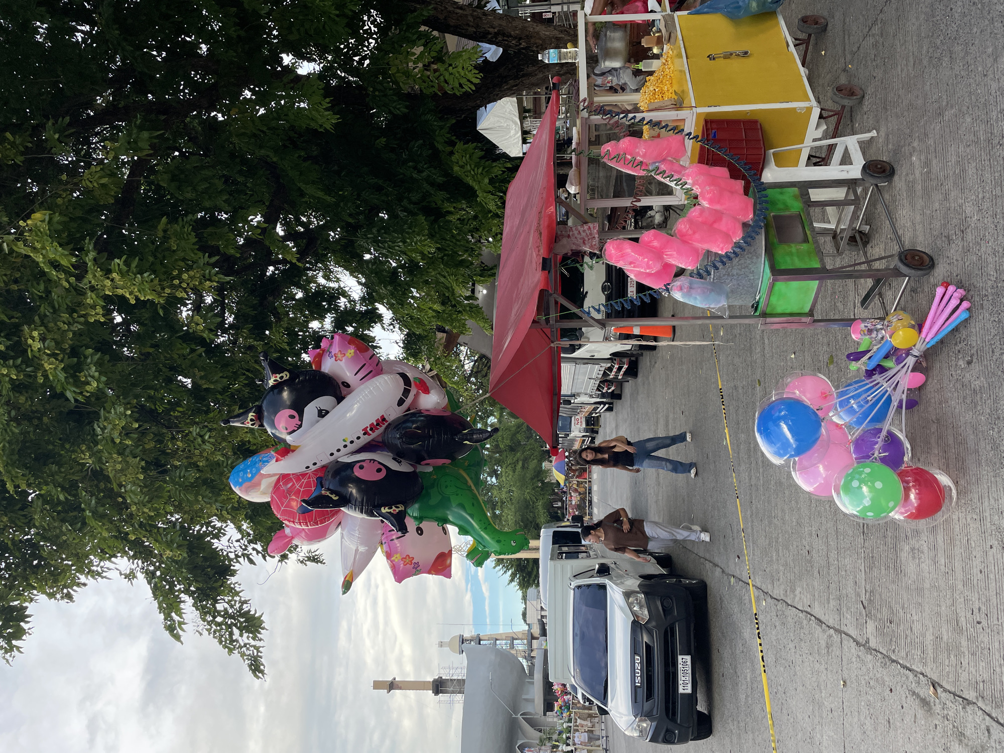 The image shows a street scene in Davao City, Philippines, likely near a public event or market. In the foreground, a collection of colorful balloons, including character-themed ones like a dinosaur, a plane, and cartoon animals, are prominently displayed. To the right, a food cart is set up, selling popcorn and cotton candy. Several people are present, including two women walking on the street near a white Isuzu truck. A yellow caution tape runs along the ground, suggesting a restricted area or ongoing activity. In the background, trees provide shade, and industrial-looking structures are visible in the distance. The overall atmosphere appears to be that of a bustling outdoor gathering.