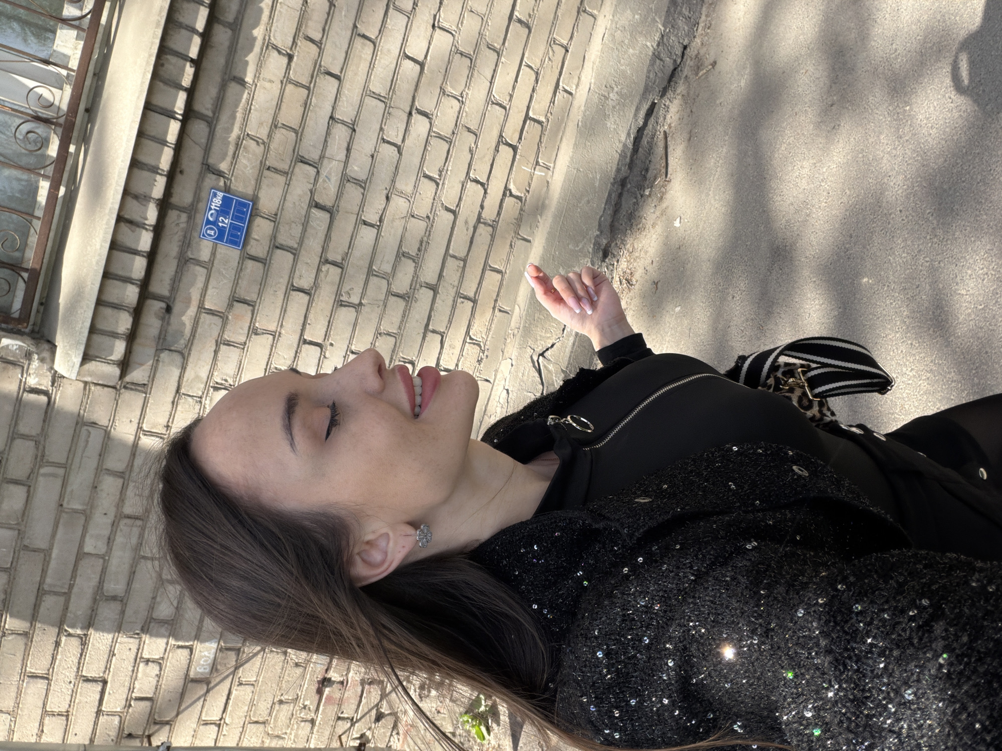 A person with long brown hair is standing outside near a brick building. The person is wearing a dark jacket with sequins. The building has a blue sign that reads "118 KG 12" on it. The location is likely in Saint Petersburg, Russia. The person has their eyes closed and is sticking their tongue out. There is a sidewalk next to the brick building.