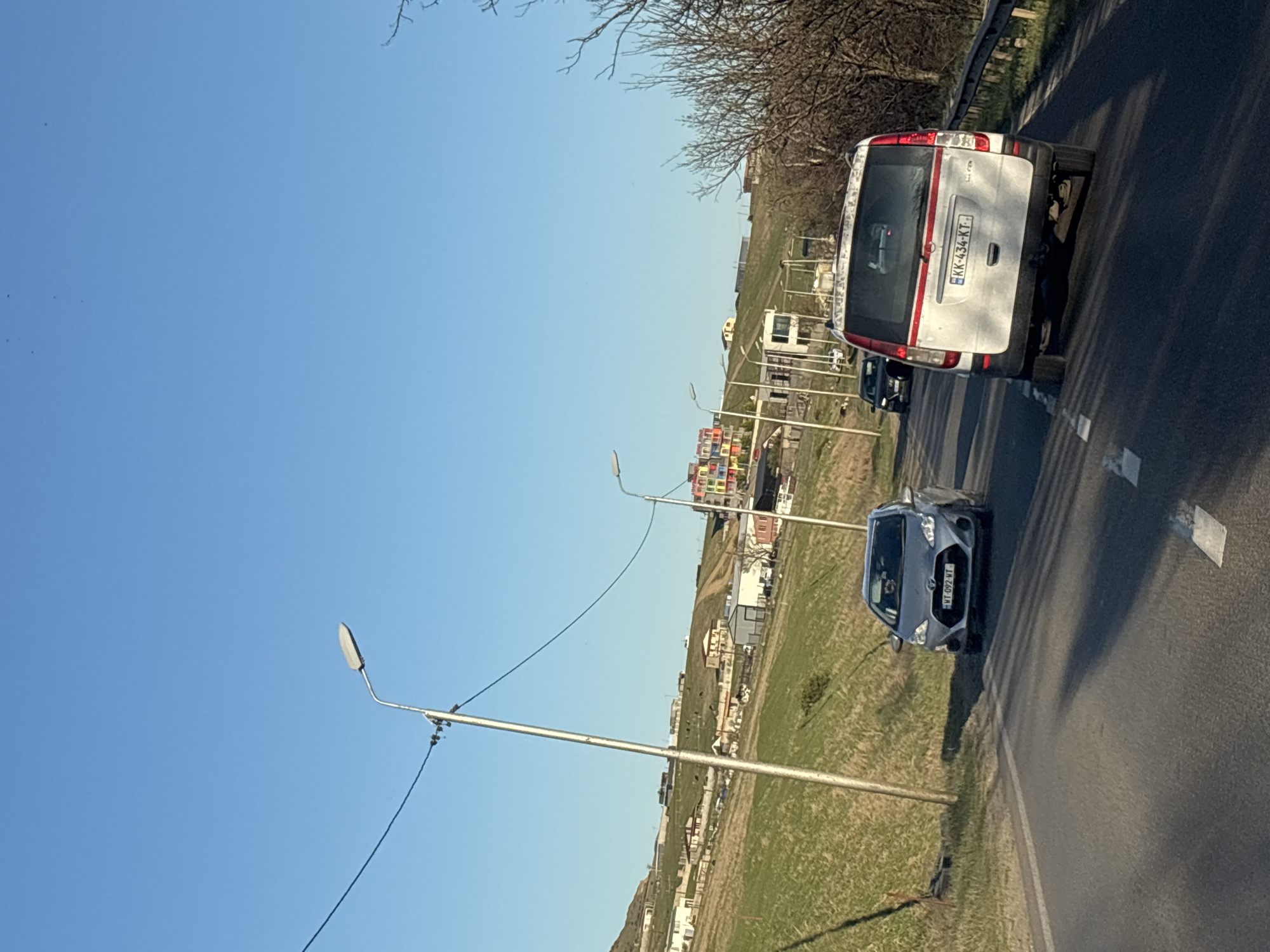 The image shows a road scene with two cars, a white van with license plate "KK-434-KT" and a gray car with license plate "WT-092-NT", on a road lined with streetlights. A grassy area is present on the side of the road with buildings in the background. The sky is clear and blue. Based on the license plates and architectural style, it is plausible this image was taken near Tbilisi, Georgia.