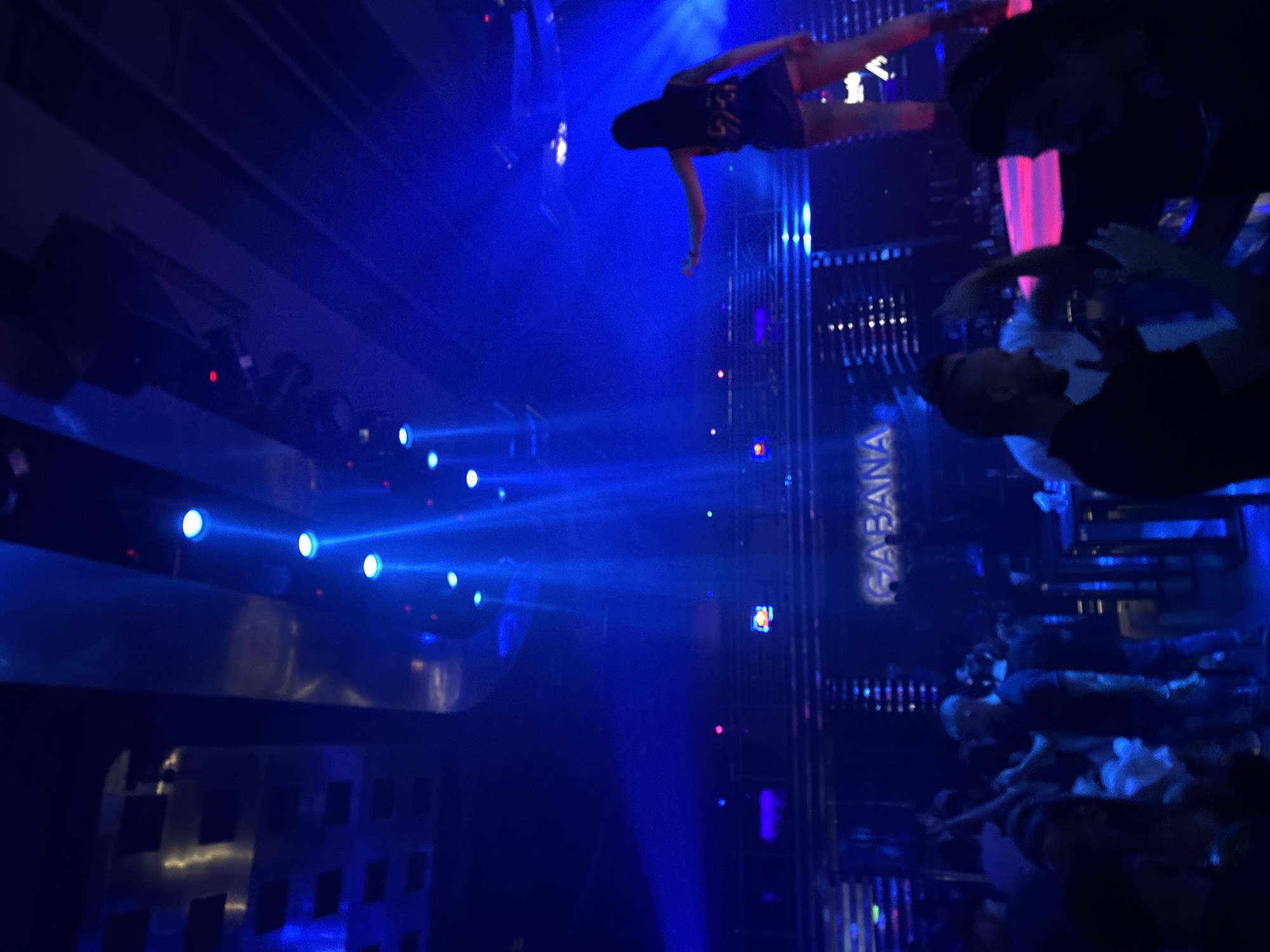This vertical image captures a vibrant and energetic scene within a dark indoor nightclub or entertainment venue in Plovdiv, Bulgaria, identified by the illuminated sign "CABANA". The dominant aesthetic is created by intense blue artificial lighting.
In the upper left, a female performer stands on an elevated platform or stage, illuminated by a bright blue spotlight. She wears a dark outfit, possibly a dress or top and shorts, and appears to be in motion with her arms outstretched, suggesting dancing or a performance. Her figure is partially silhouetted against the bright blue light and hazy air.
To the right and below the performer, several people, mostly men, are visible, likely members of the audience or other attendees. Their forms are somewhat obscured by the low light and motion, but some appear to be looking towards the stage. One person is discernible in a white shirt.
Numerous powerful blue spotlights project visible beams through what appears to be smoke or haze, creating a dynamic and immersive atmosphere. Other small, colored lights (red, pink, purple) are scattered throughout the background, adding to the club ambiance. Structural elements of the venue, including dark horizontal beams and vertical panels, are also visible. The overall scene suggests a lively night event, such as a concert or a club night, with a performer engaging an audience in a vibrant, light-filled environment.