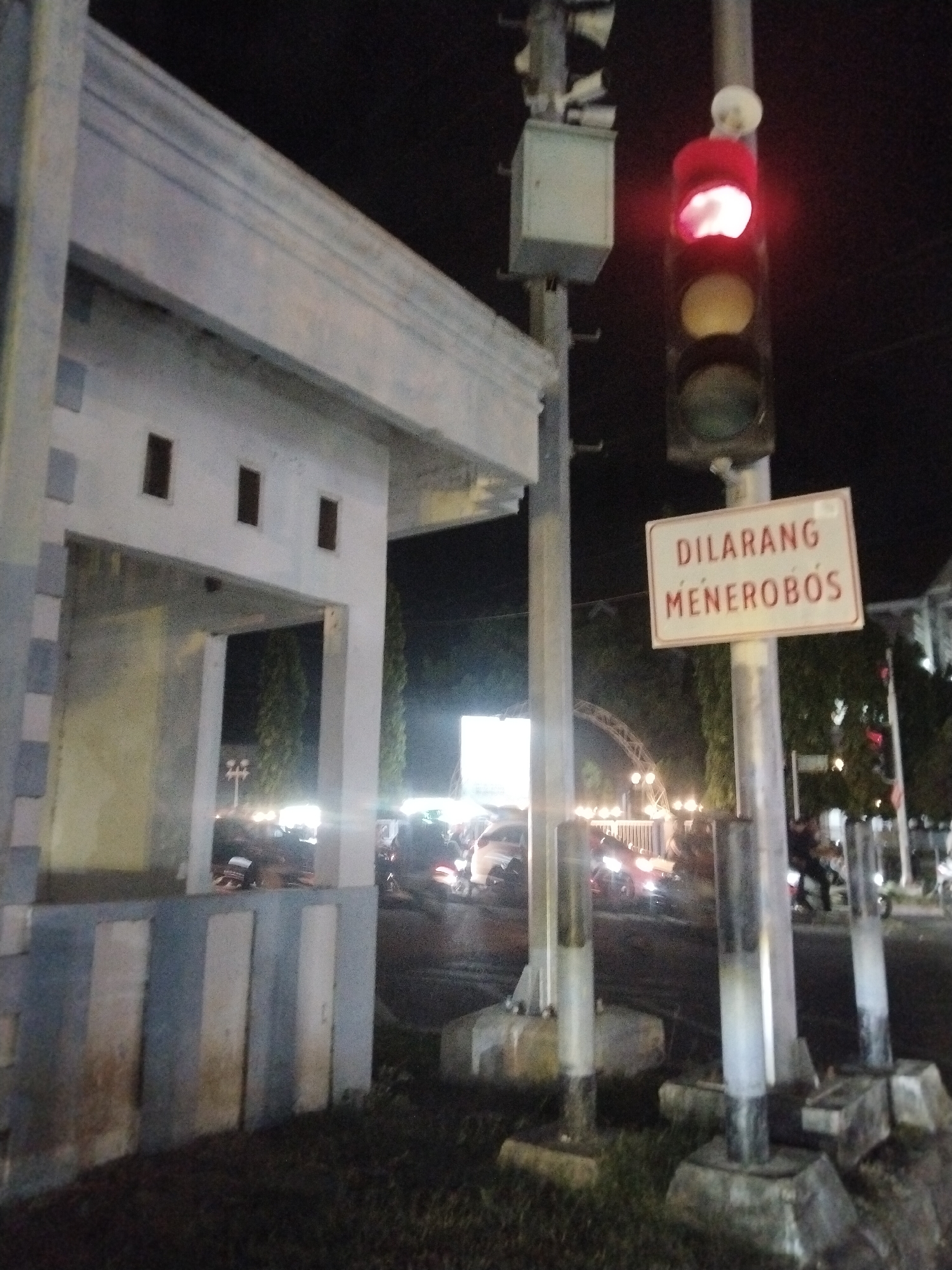 The image depicts a street scene at night in Lhokseumawe, Indonesia. A traffic light shows red with a sign that reads "DILARANG MENEROBOS", meaning "DO NOT RUN". In the background, a building with an arched entrance is illuminated by lights. Cars and motorcycles are present on the road. There's a structure with a geometric facade to the left, partially visible. The area surrounding the traffic signal pole has patches of grass.