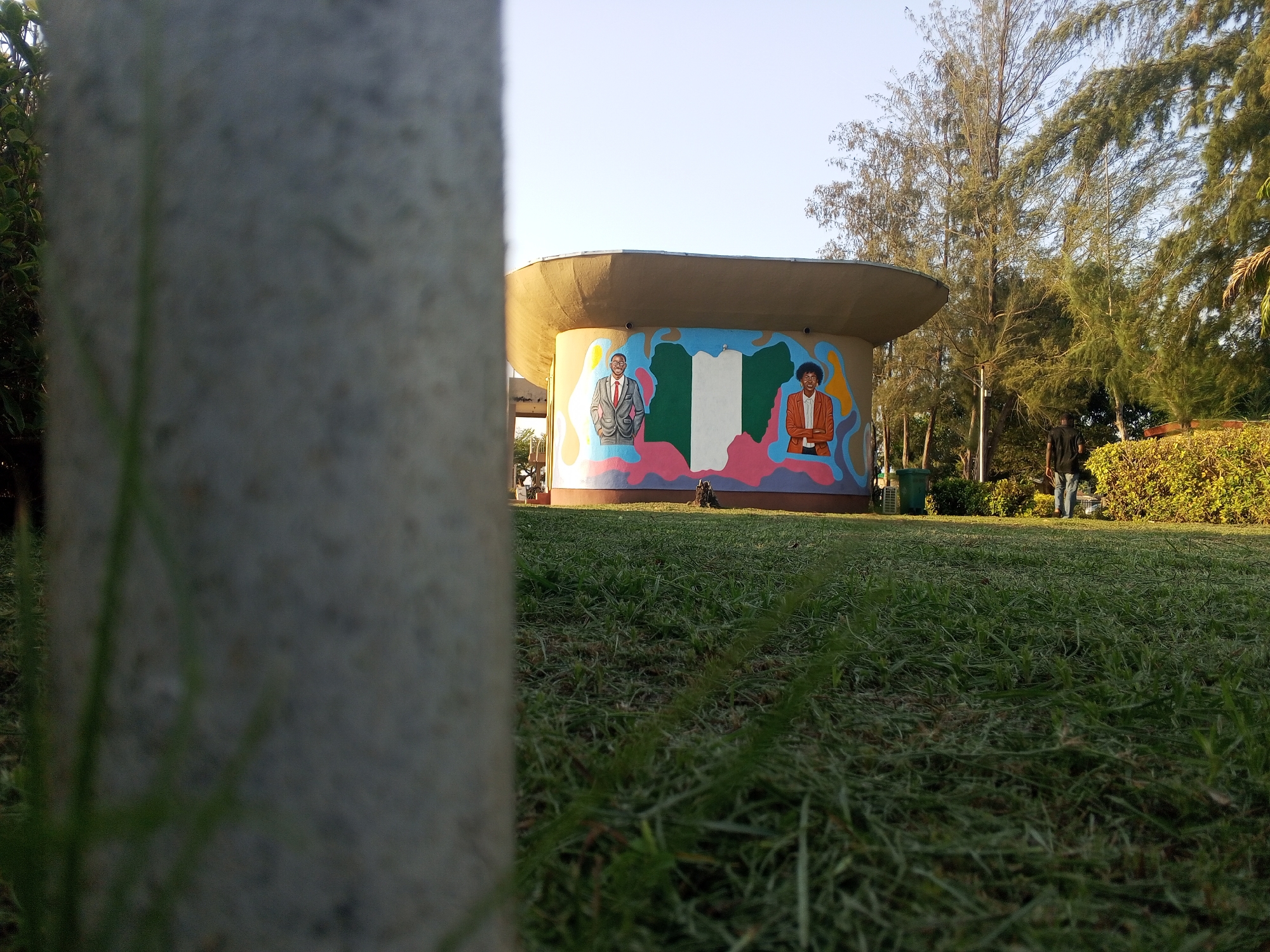 The image is taken from a low angle in a park in Tawpo, Nigeria. A blurred grey concrete pillar dominates the foreground. Beyond the pillar is a green lawn, and further back is a cylindrical building with a flat roof. The building has a mural depicting two people and the Nigerian flag. Trees and shrubs surround the building. The sky is light blue.