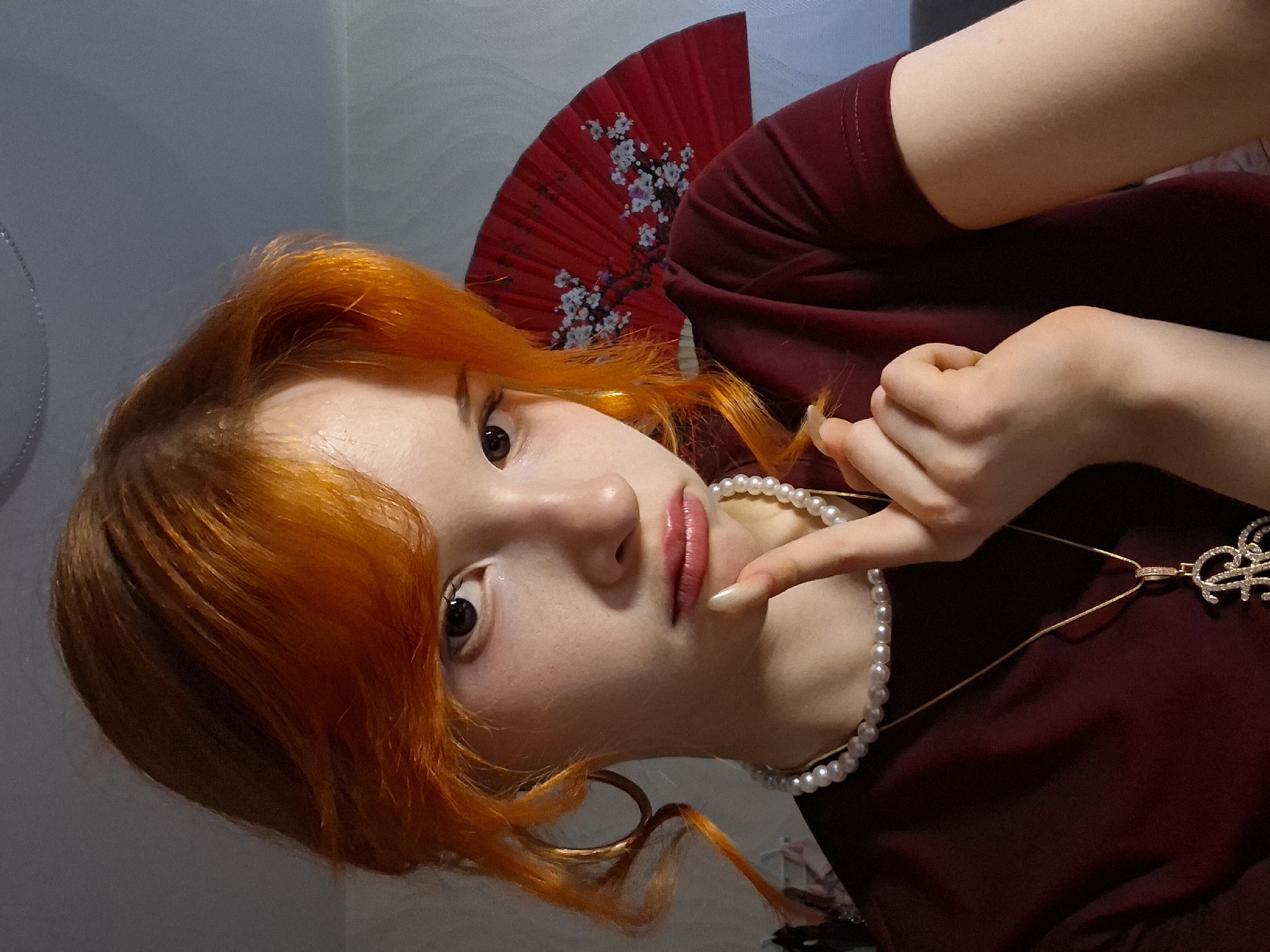 A young woman with orange hair and fair skin is wearing a maroon top, a pearl necklace, and a gold necklace with a sparkly pendant. Her index finger is resting on her chin. Behind her is a red fan with a white floral design. The background is a muted gray. The image appears to be taken indoors, possibly in a residential setting. There is no specific information present that indicates the location is Chaikovsky, Russia.